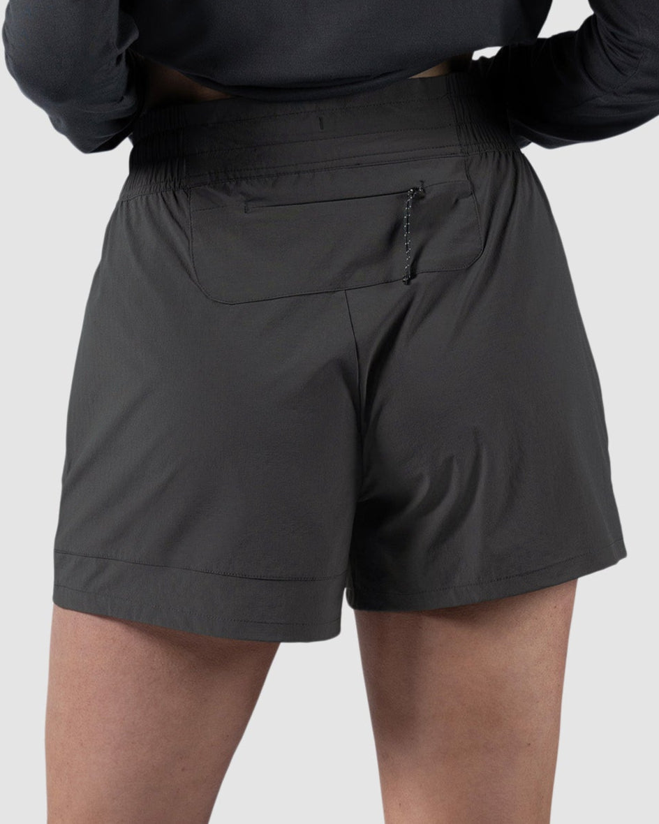 A person stands facing away against a plain light gray background, wearing a long-sleeve black shirt, white sneakers, and BEARDEDGOAT Womens Alpina Short—high-waisted adventure shorts with zippered pockets.
