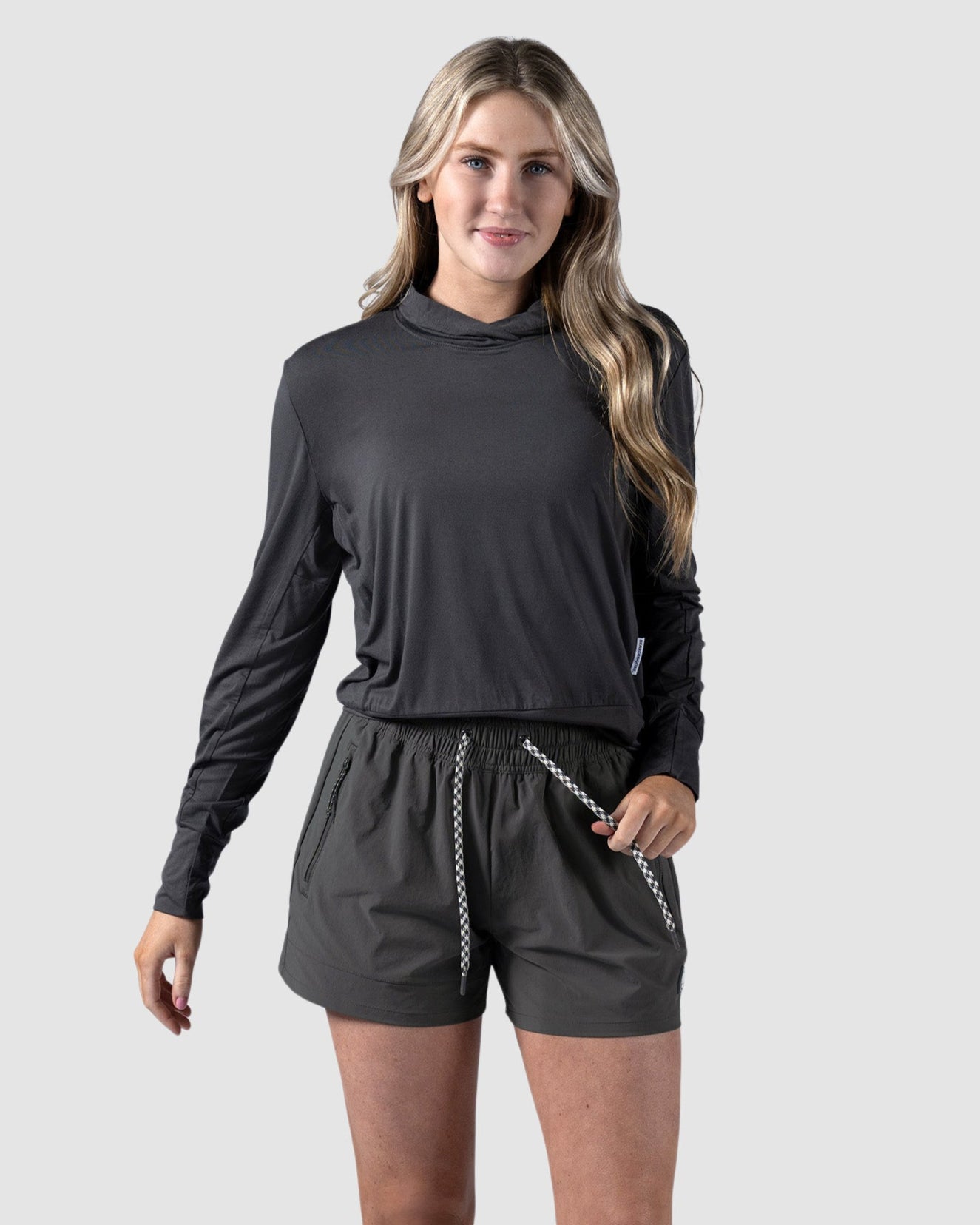 A woman with long wavy hair stands against a light gray background, smiling slightly, wearing BEARDEDGOATs Womens Alpina Short—high-waisted adventure shorts with zippered pockets—a dark gray long-sleeve hoodie, and white sneakers. One hand is in her pocket.