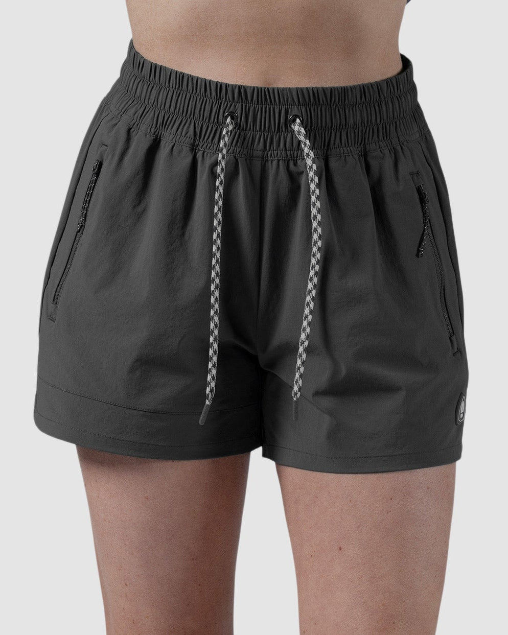 A person wears BEARDEDGOAT Womens Alpina Short in dark gray stretchy ripstop with drawstring and zippered pockets, paired with white sneakers and a bare midriff, standing against a plain light gray background.