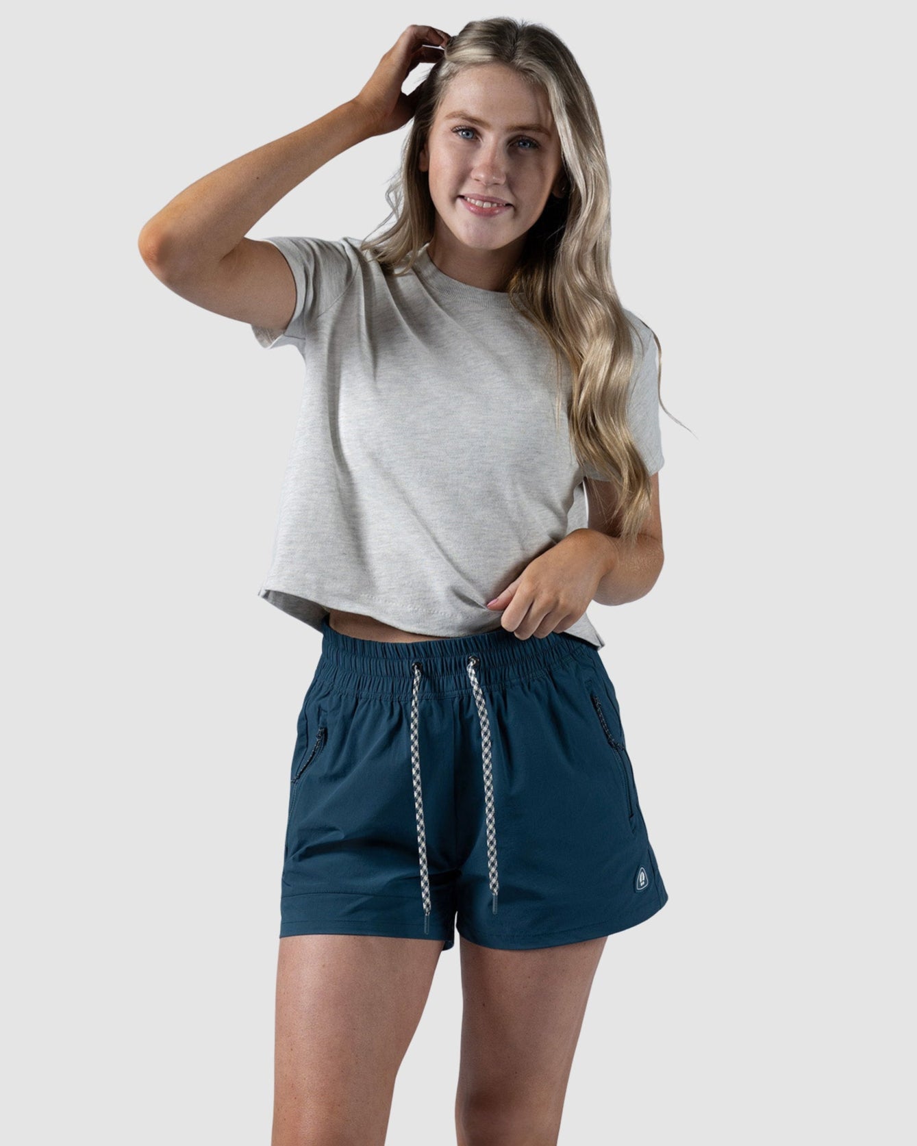 A woman wearing a light gray tee, BEARDEDGOAT Womens Alpina Short in dark blue with zippered pockets, and white sneakers stands against a plain light background, smiling and touching her hair.