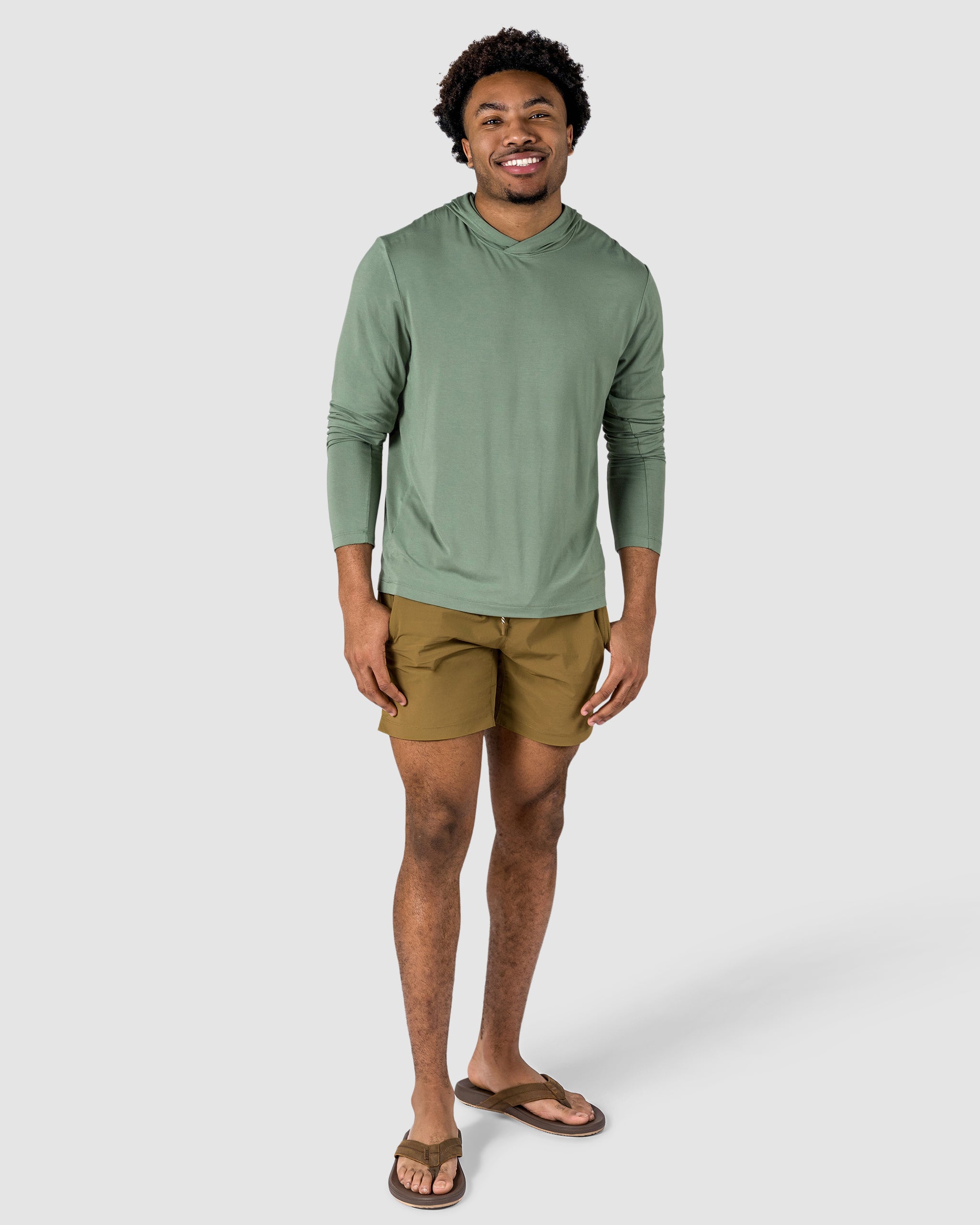 A smiling man in a light green long-sleeve hoodie, BEARDEDGOAT Mens Rambler Short, and flip-flops stands against a plain light gray background.