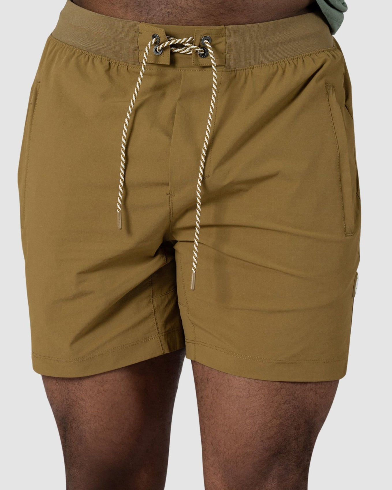 A person wearing BEARDEDGOAT Mens Rambler Short in mustard yellow, featuring 4-way stretch, and part of a green shirt stands against a plain light background. Only the lower torso and upper legs are visible.