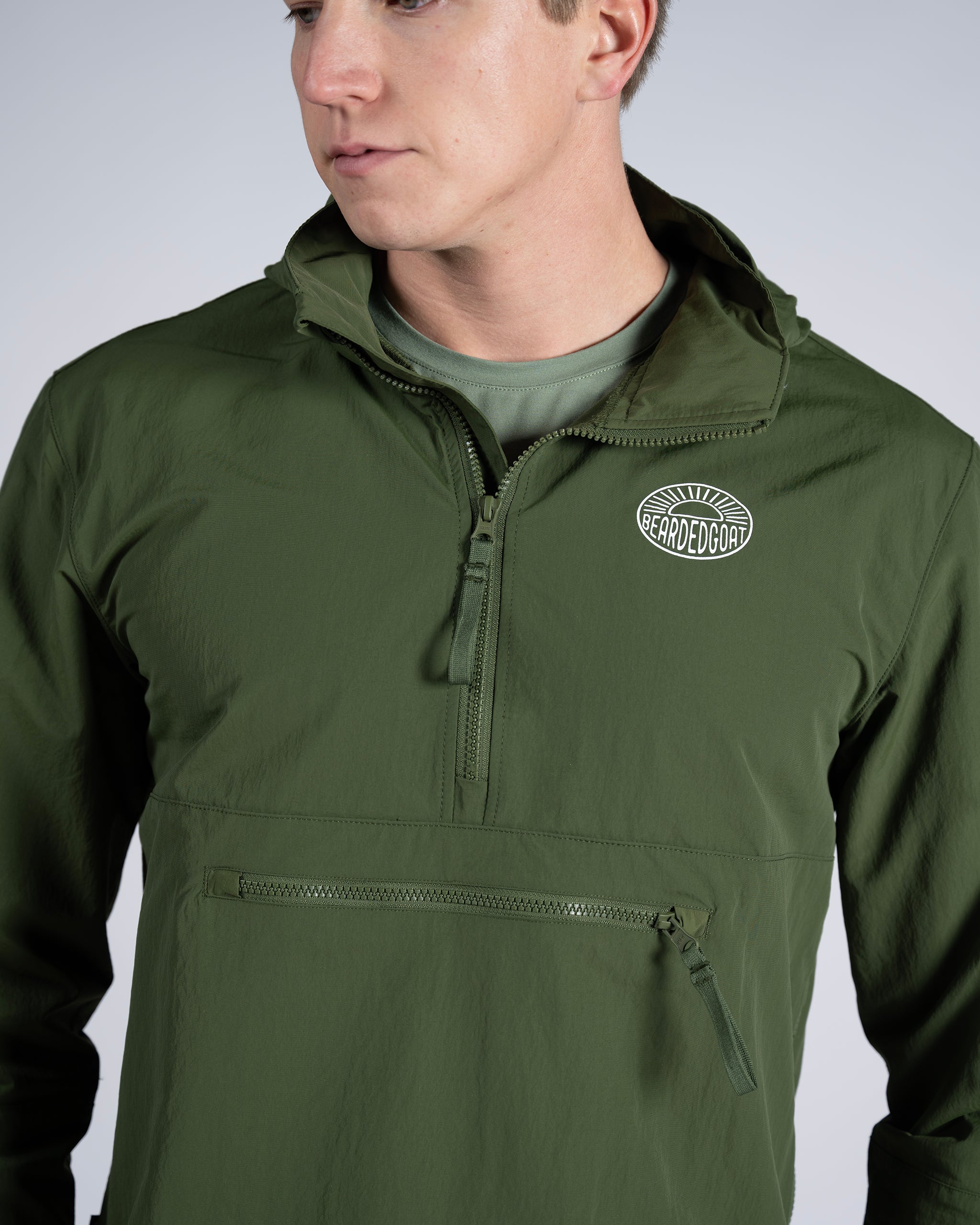 A man models the BEARDEDGOAT Mens Deluge Pullover in olive green, featuring a lightweight design, zippered chest pocket, and Bearded Goat patch logo on the left side, while looking to the side against a plain background.