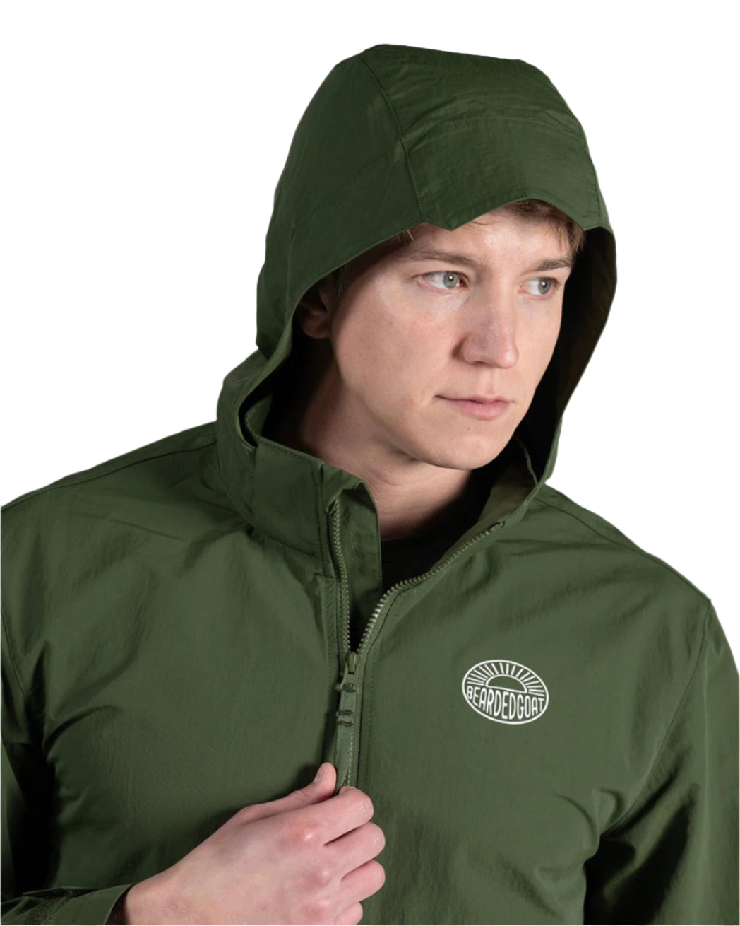 A man wearing the BEARDEDGOAT Mens Deluge Pullover—an olive green, lightweight hooded jacket with a chest logo and zipper—stands against a light gray background, looking right and holding the collar.