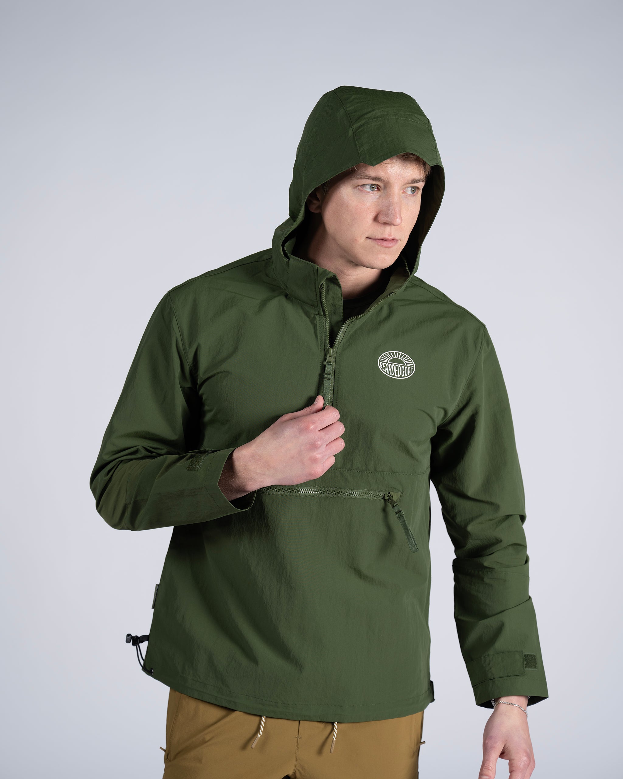 A man wearing the BEARDEDGOAT Mens Deluge Pullover—an olive green, lightweight hooded jacket with a chest logo and zipper—stands against a light gray background, looking right and holding the collar.