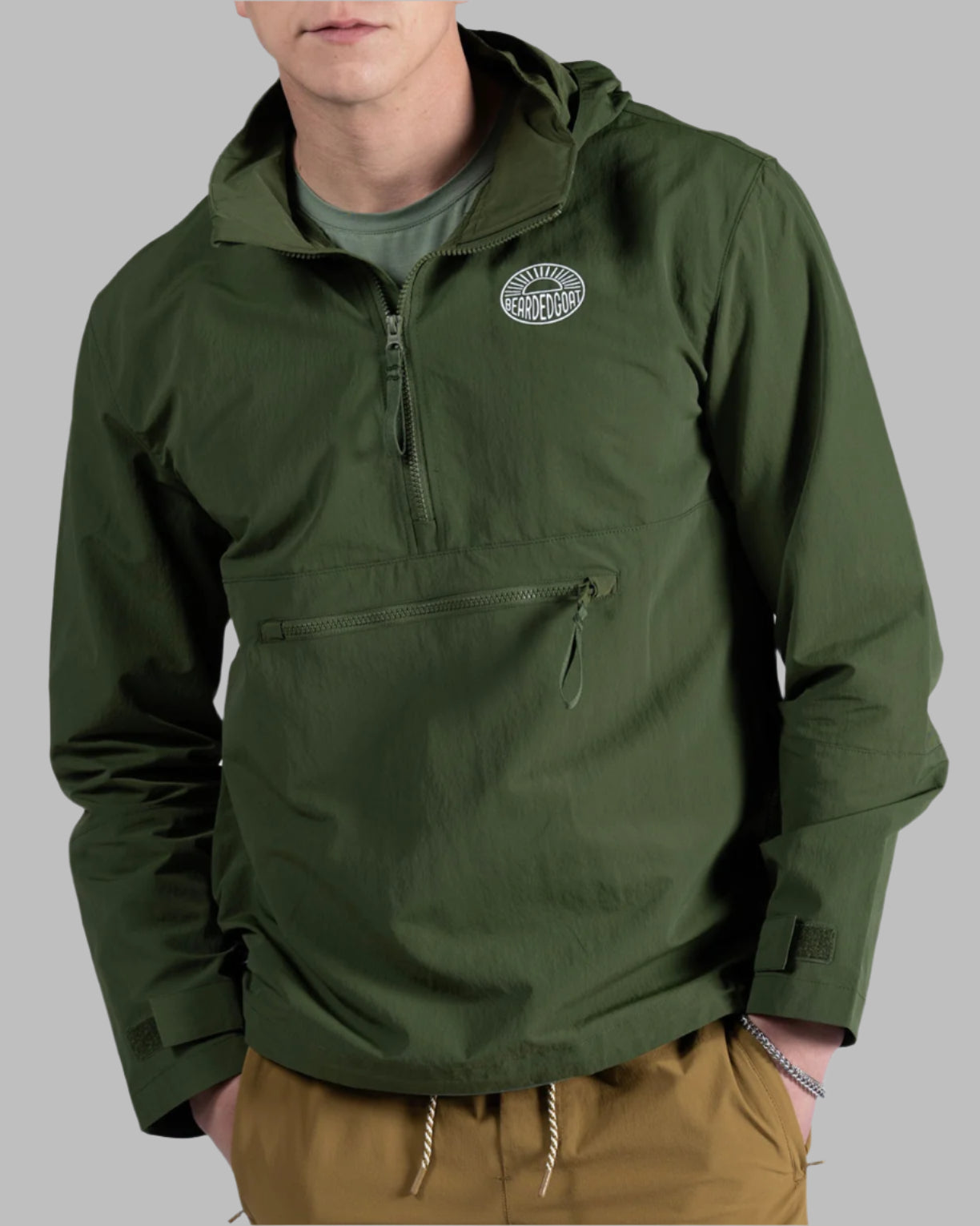 A young man stands against a plain background, wearing the BEARDEDGOAT Mens Deluge Pullover—a green hooded jacket with chest logo and lightweight packable design—paired with a green t-shirt and tan shorts, hands in pockets, looking at the camera.