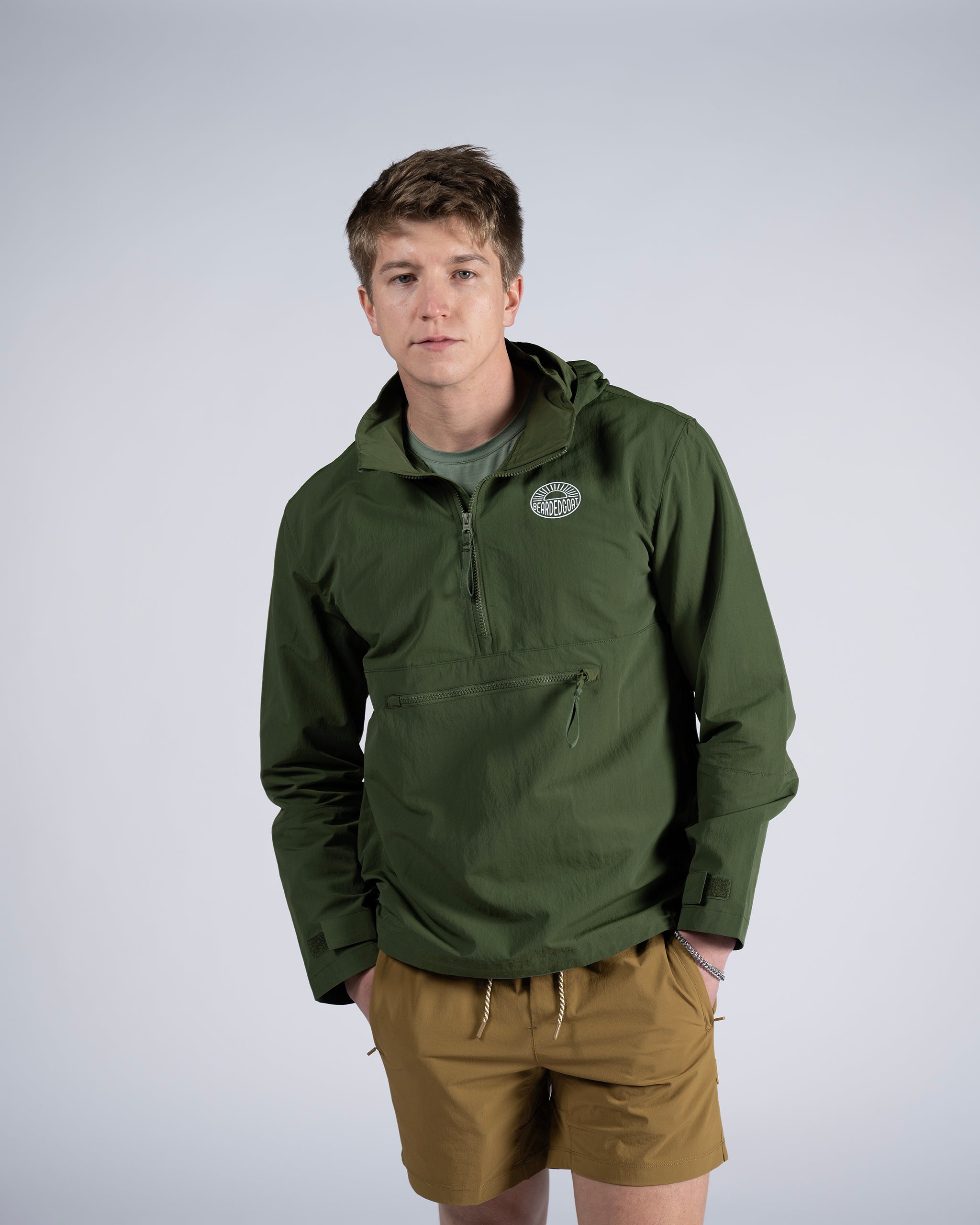 A young man stands against a plain background, wearing the BEARDEDGOAT Mens Deluge Pullover—a green hooded jacket with chest logo and lightweight packable design—paired with a green t-shirt and tan shorts, hands in pockets, looking at the camera.