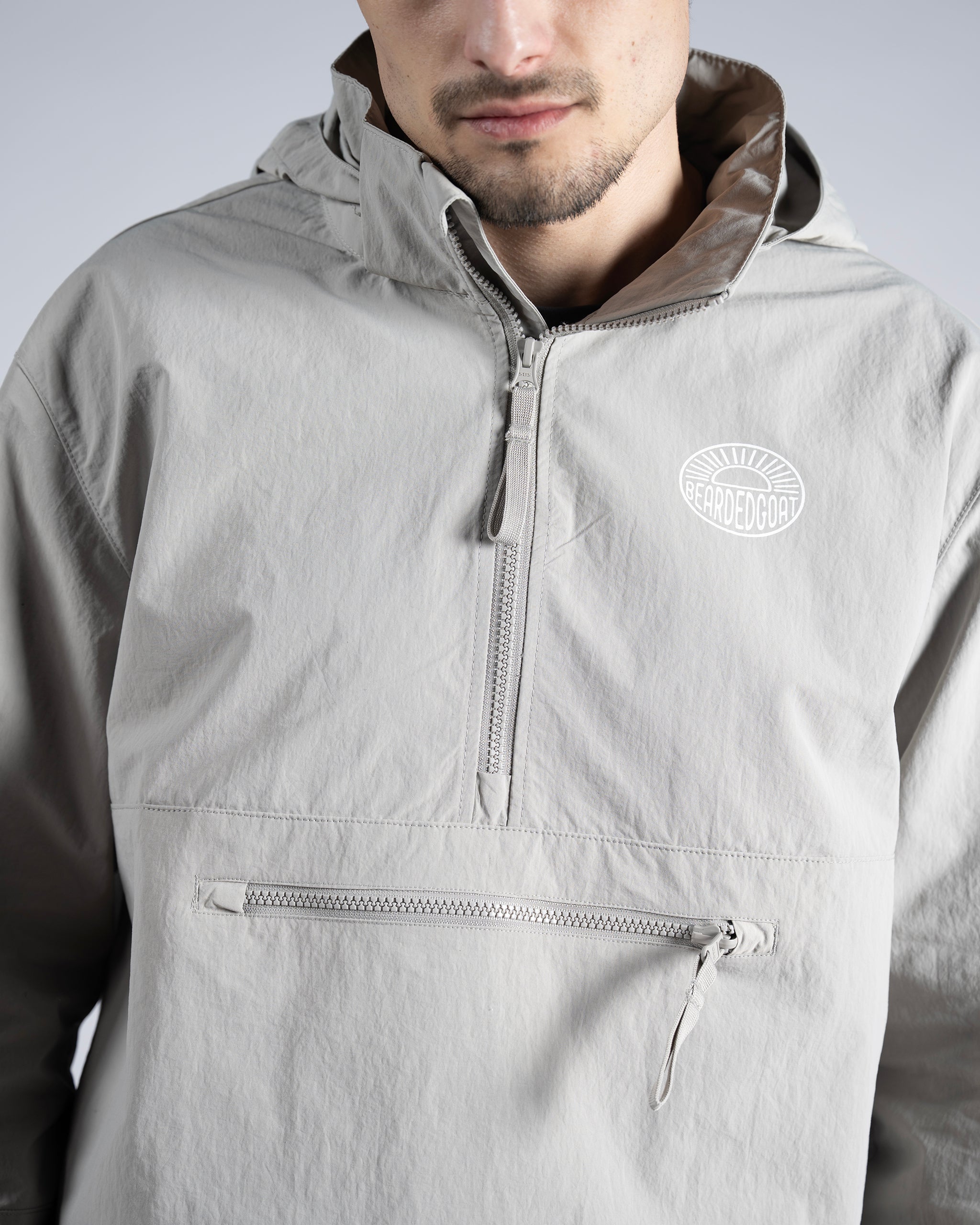 A man wears the BEARDEDGOAT Mens Deluge Pullover, a lightweight light gray hooded jacket with a half-zip front and zippered pocket. The BEARDEDGOAT logo appears on the left chest. The photo shows him from shoulders to mid-torso.