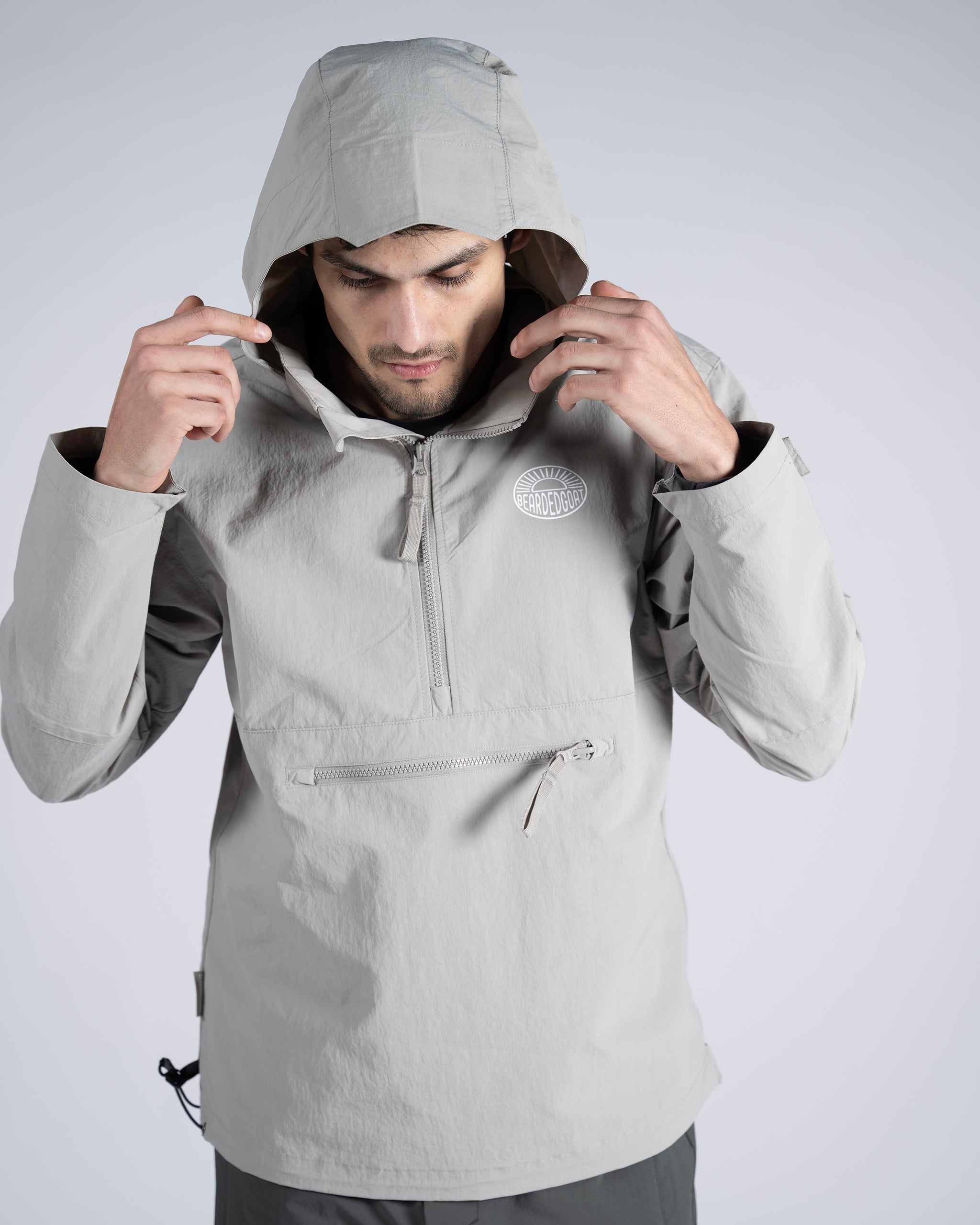 A man wearing the BEARDEDGOAT Mens Deluge Pullover, a lightweight light gray hooded jacket with a front zipper and chest pocket, stands against a plain background, looking down as he adjusts his hood with both hands.
