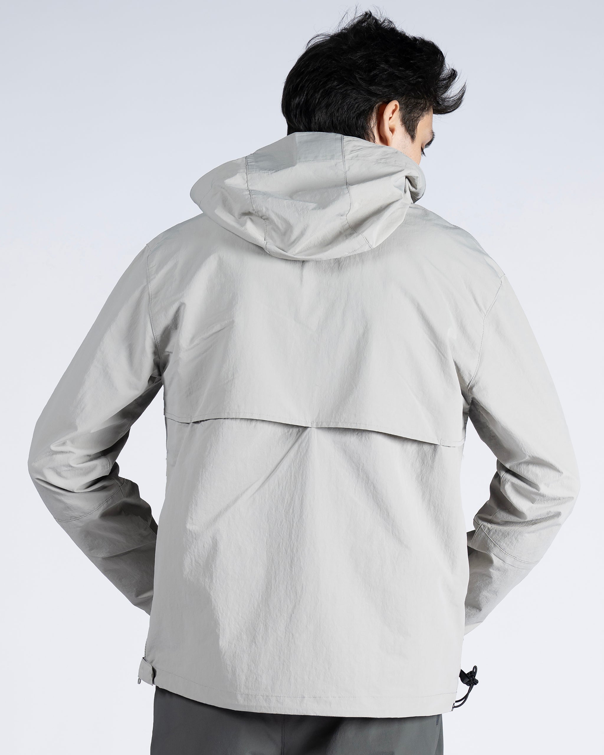 A person wears the BEARDEDGOAT Mens Deluge Pullover, a light gray, water-resistant jacket with a hood, vented back, and long sleeves. The hands-in-pockets stance showcases its lightweight, packable design for on-the-go comfort.