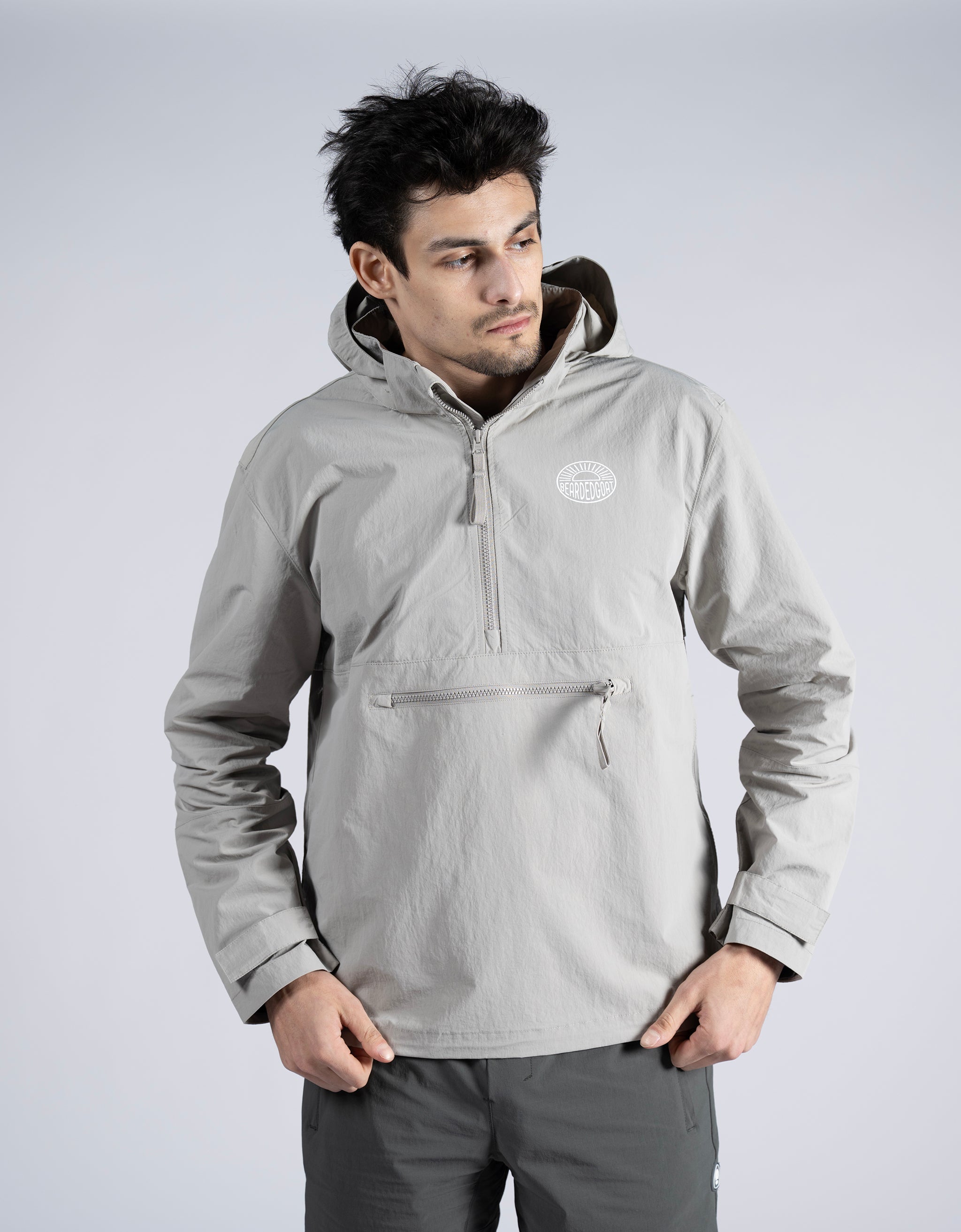A man with dark hair models the BEARDEDGOAT Mens Deluge Pullover—a lightweight, light gray hooded jacket with a front zipper pocket—paired with matching gray pants, standing before a plain light gray background.