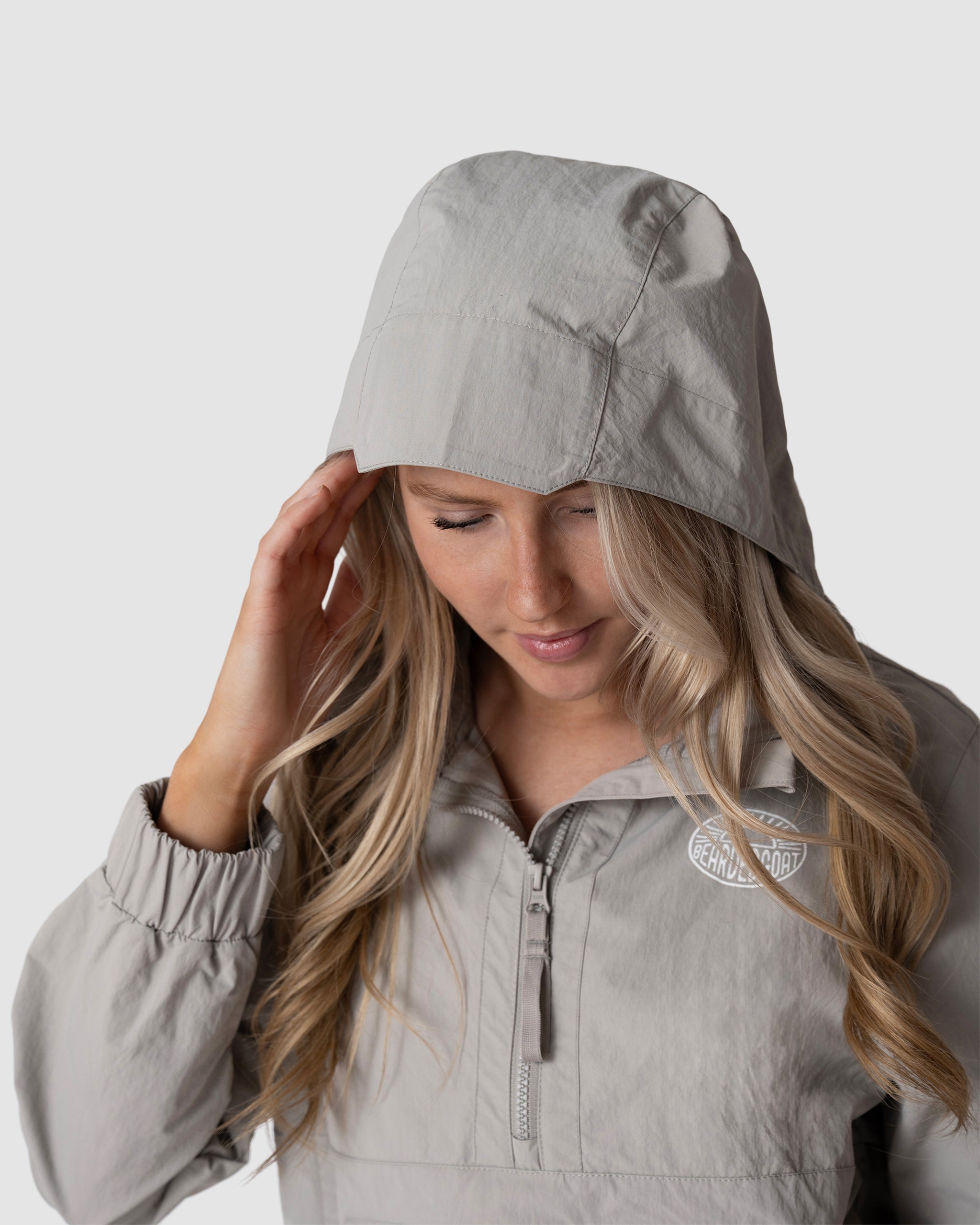 A woman with long blonde hair wears the BEARDEDGOAT Womens Deluge Pullover, a light gray water-resistant jacket with a removable hood. She looks down, touching the hood, against a plain light gray background.