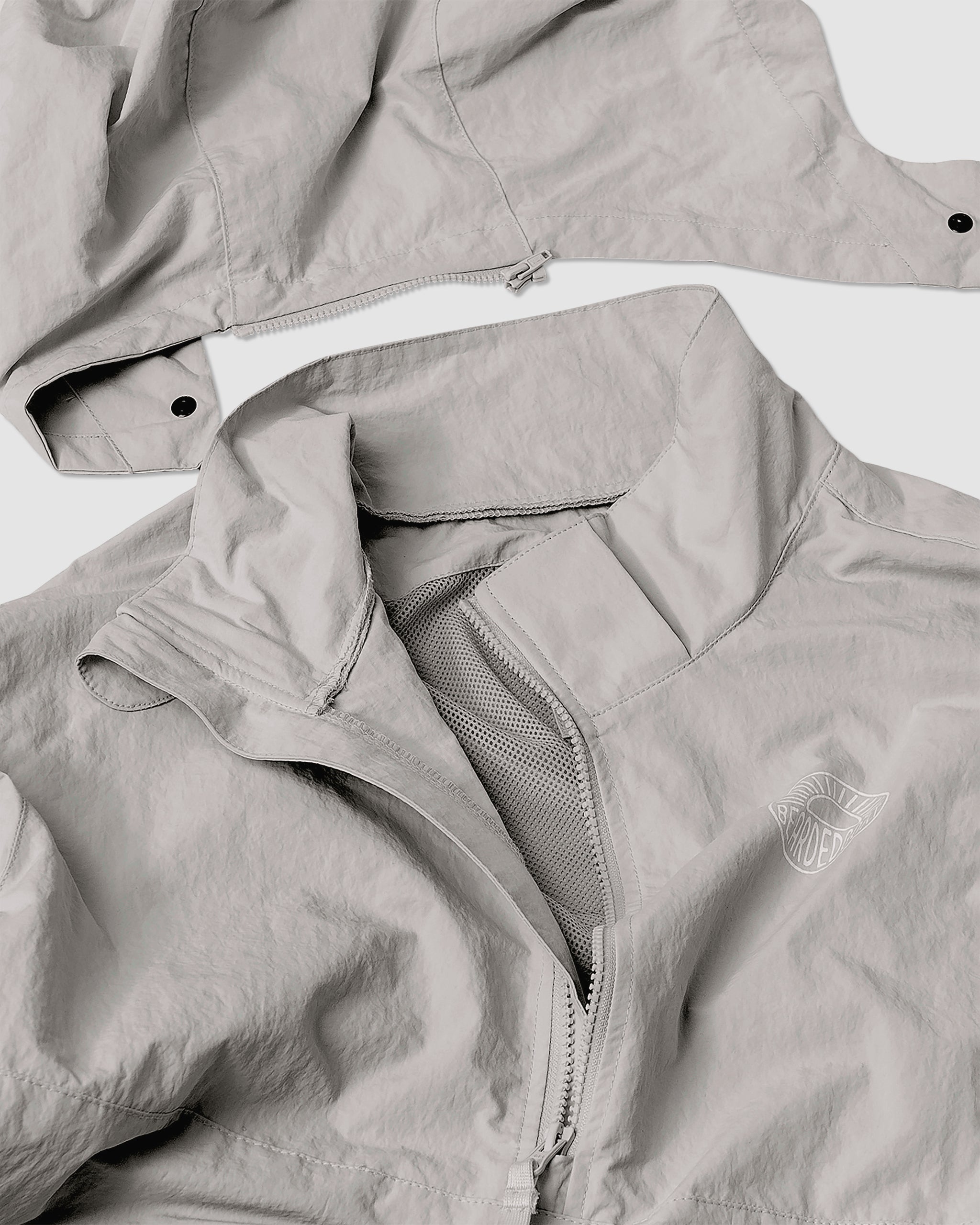 The BEARDEDGOAT Womens Deluge Pullover is a light gray, water-resistant jacket with a zippered front, mesh lining, detachable shoulder section, and a small white embroidered chest logo for a sleek look.