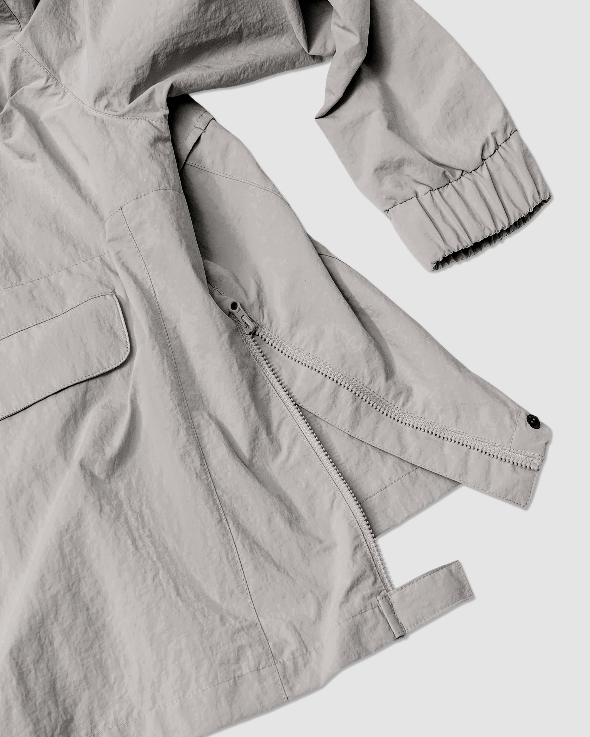 The BEARDEDGOAT Womens Deluge Pullover in light gray features a partially unzipped diagonal zipper, a flap pocket, and an elastic cuff on the sleeve, shown against a plain light background.