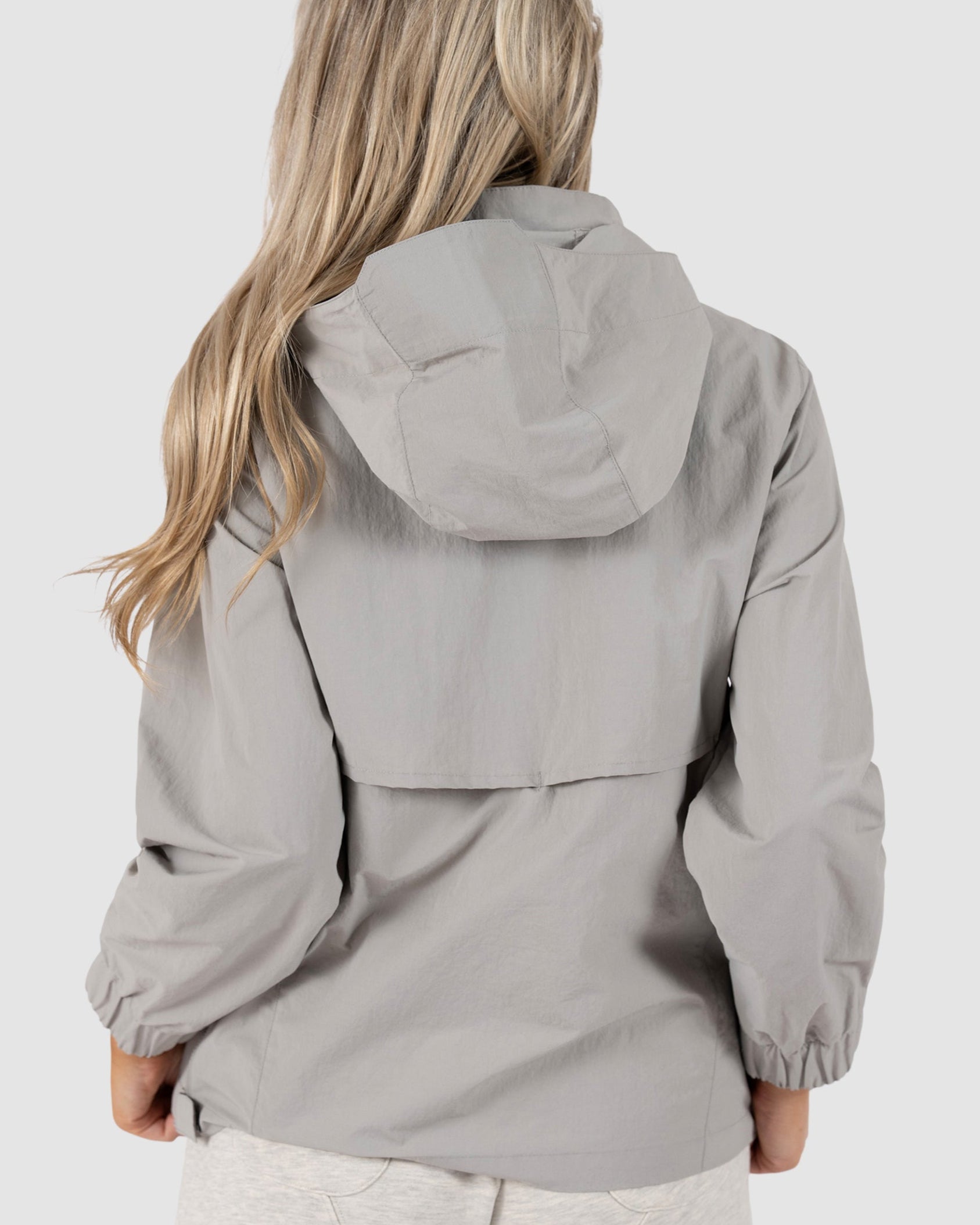 A woman with long, wavy blonde hair is seen from behind wearing the BEARDEDGOAT Womens Deluge Pullover, a light gray water-resistant jacket with removable hood, and light shorts against a plain light gray background.