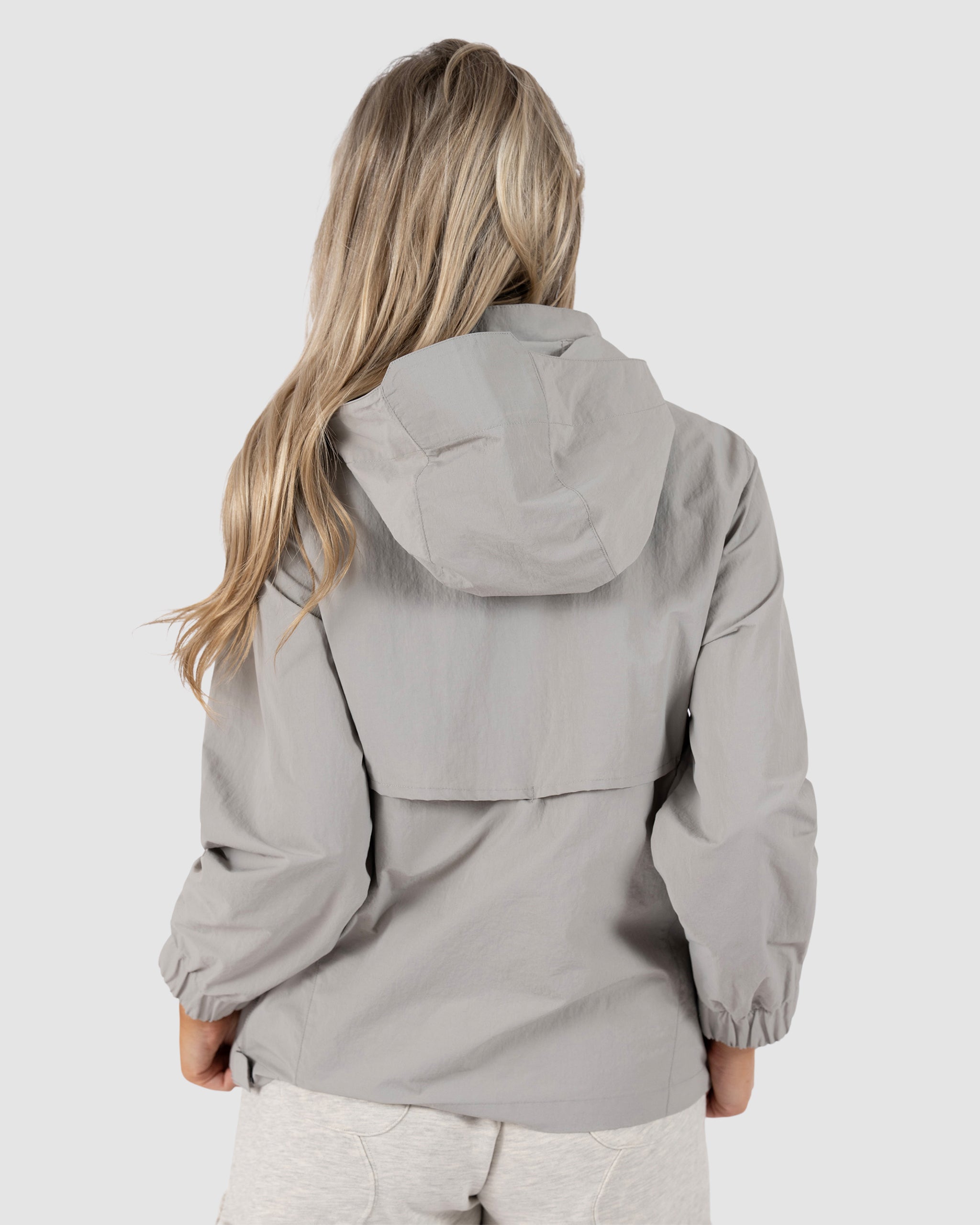A woman with long, wavy blonde hair is seen from behind wearing the BEARDEDGOAT Womens Deluge Pullover, a light gray water-resistant jacket with removable hood, and light shorts against a plain light gray background.