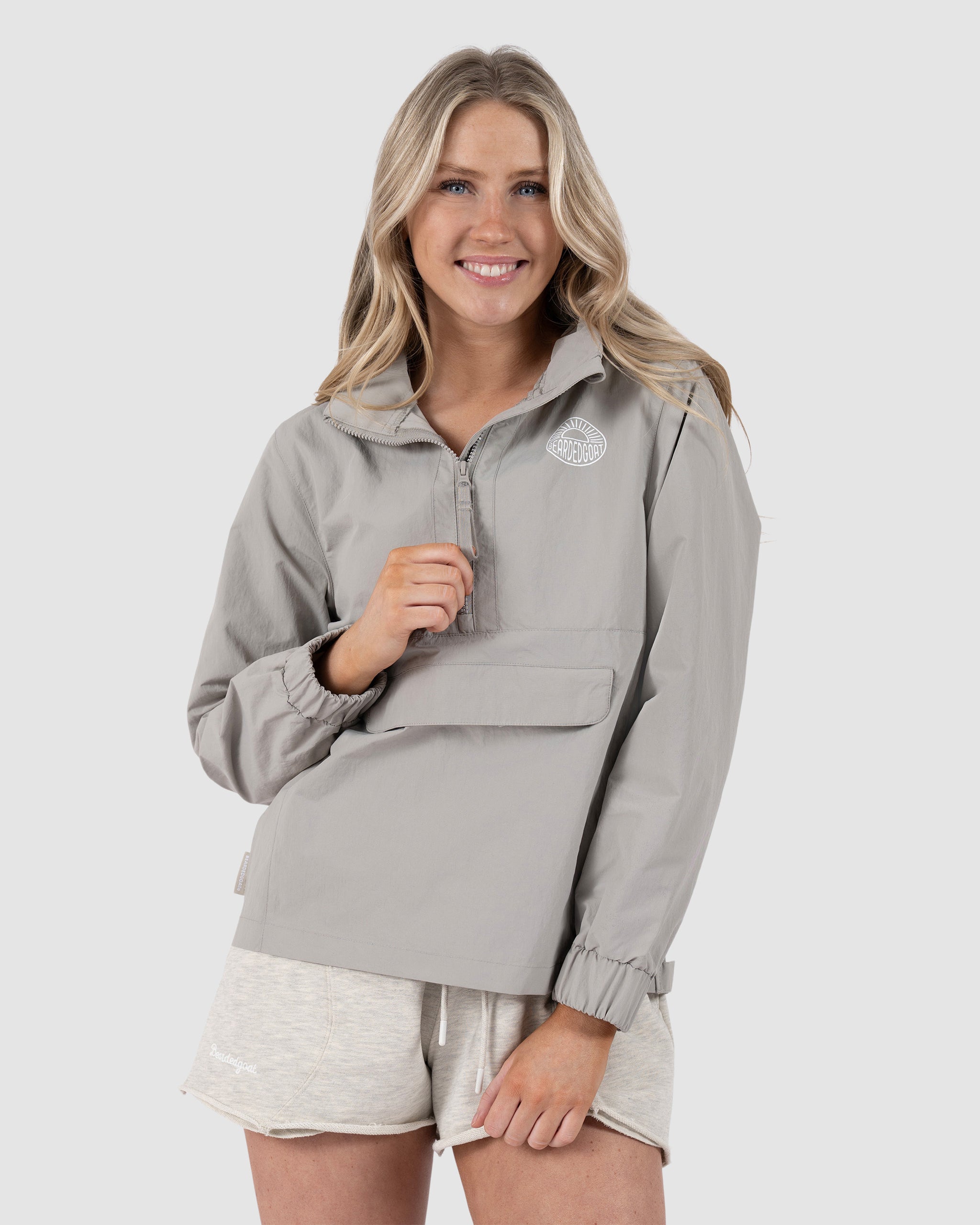 A woman with long blonde hair smiles while wearing the BEARDEDGOAT Womens Deluge Pullover in light gray, featuring water resistance and a removable hood, paired with light shorts against a plain background.