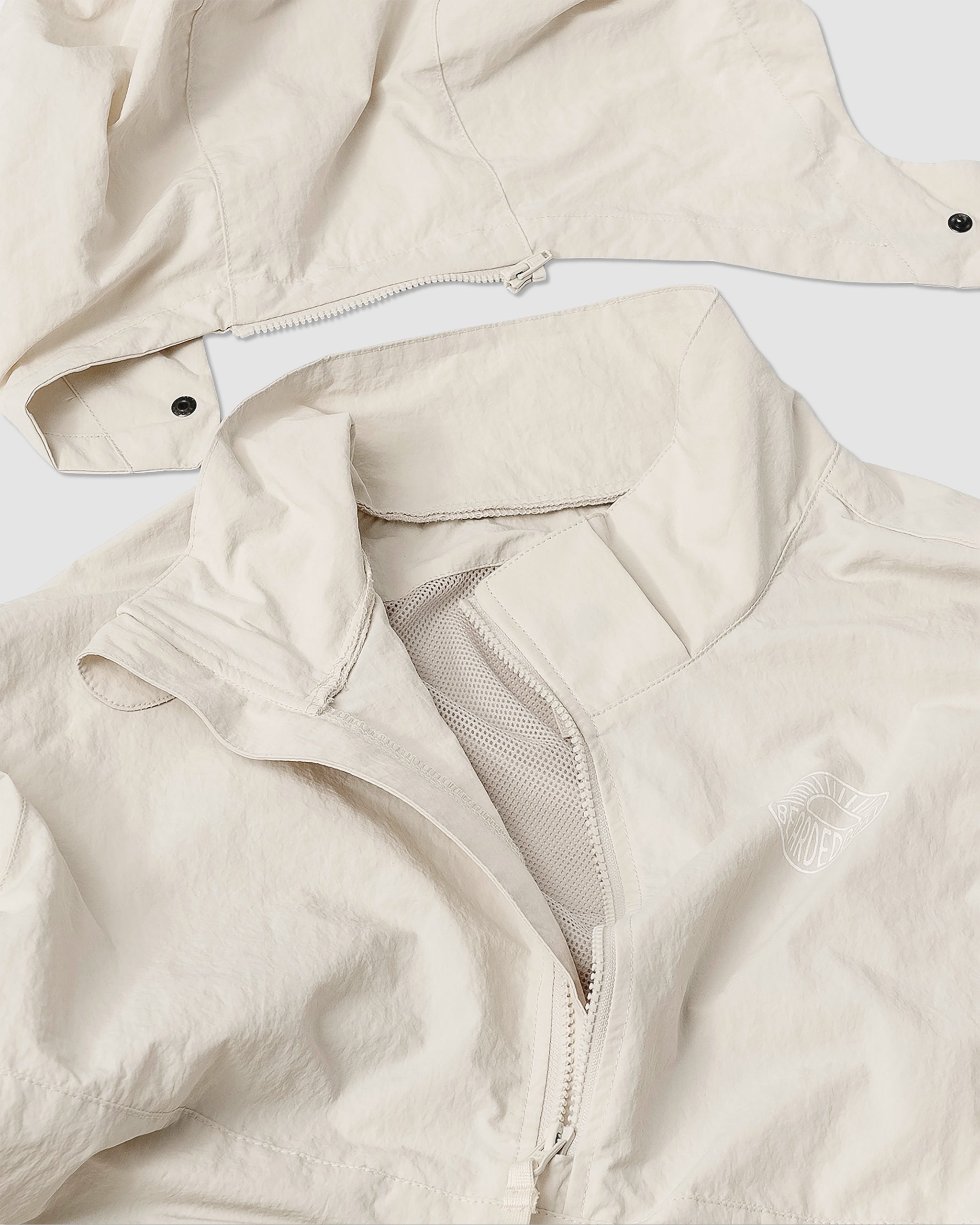 A close-up of the BEARDEDGOAT Womens Deluge Pullover, a light beige, water-resistant jacket with mesh lining, partially unzipped front, and removable hood featuring snap buttons and a zipper, laid flat on a white background.