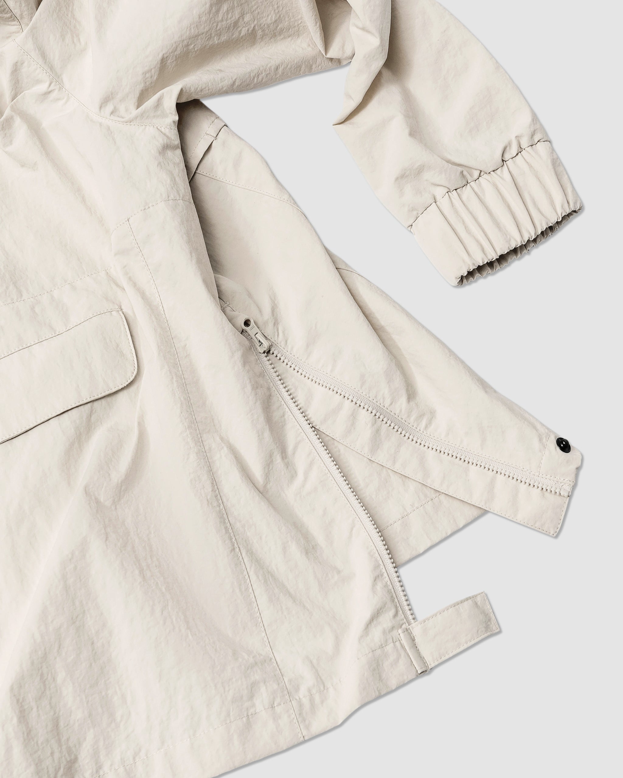 A close-up of the BEARDEDGOAT Womens Deluge Pullover in light beige shows a side zipper, flap pocket, adjustable strap detail, and elastic cuffs. The water-resistant fabric has a slightly crinkled texture.