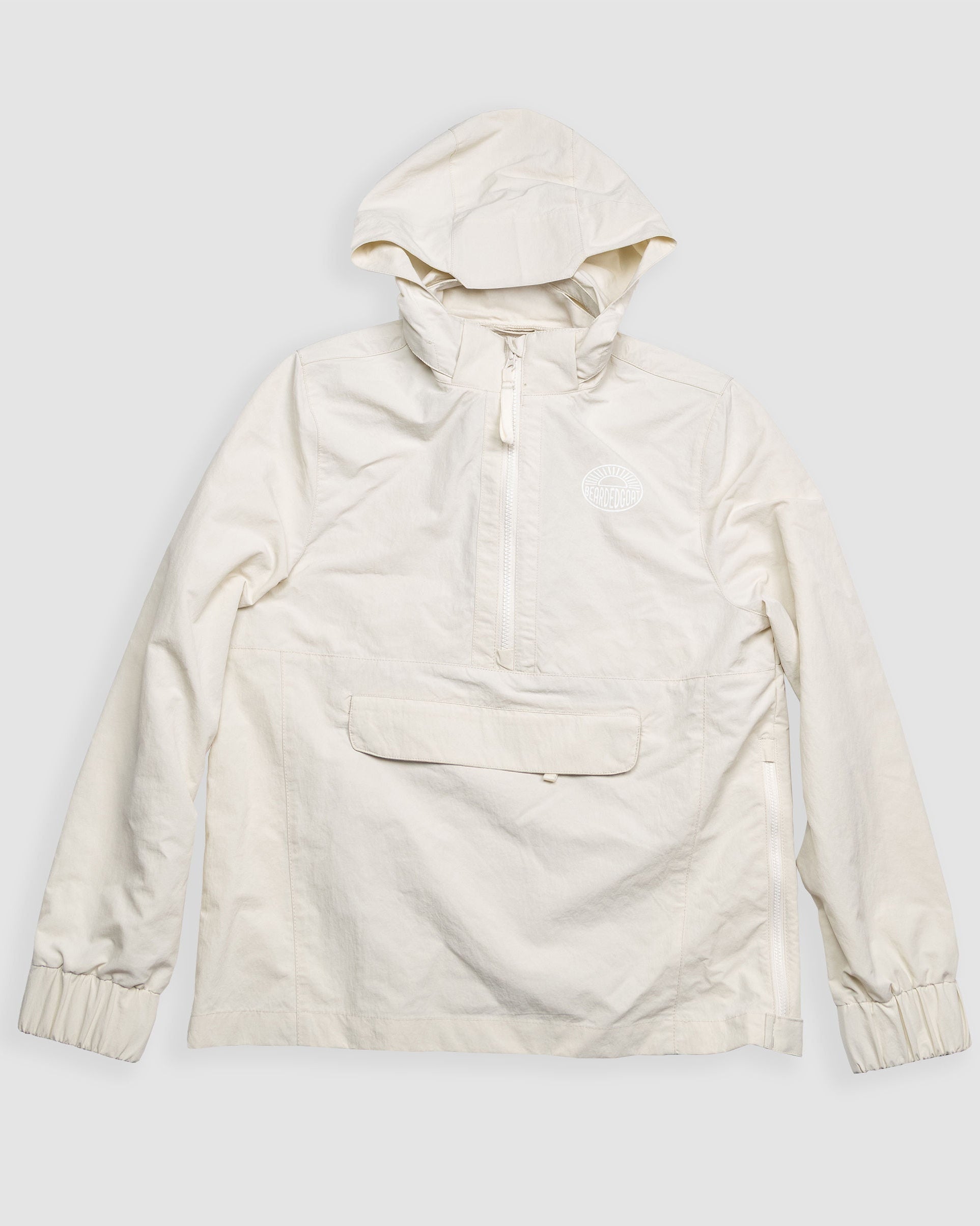 The BEARDEDGOAT Womens Deluge Pullover is a light beige, water-resistant hooded jacket with a front zipper, large flap pocket, elastic cuffs, and small circular chest logo, shown laid flat on a plain white background.