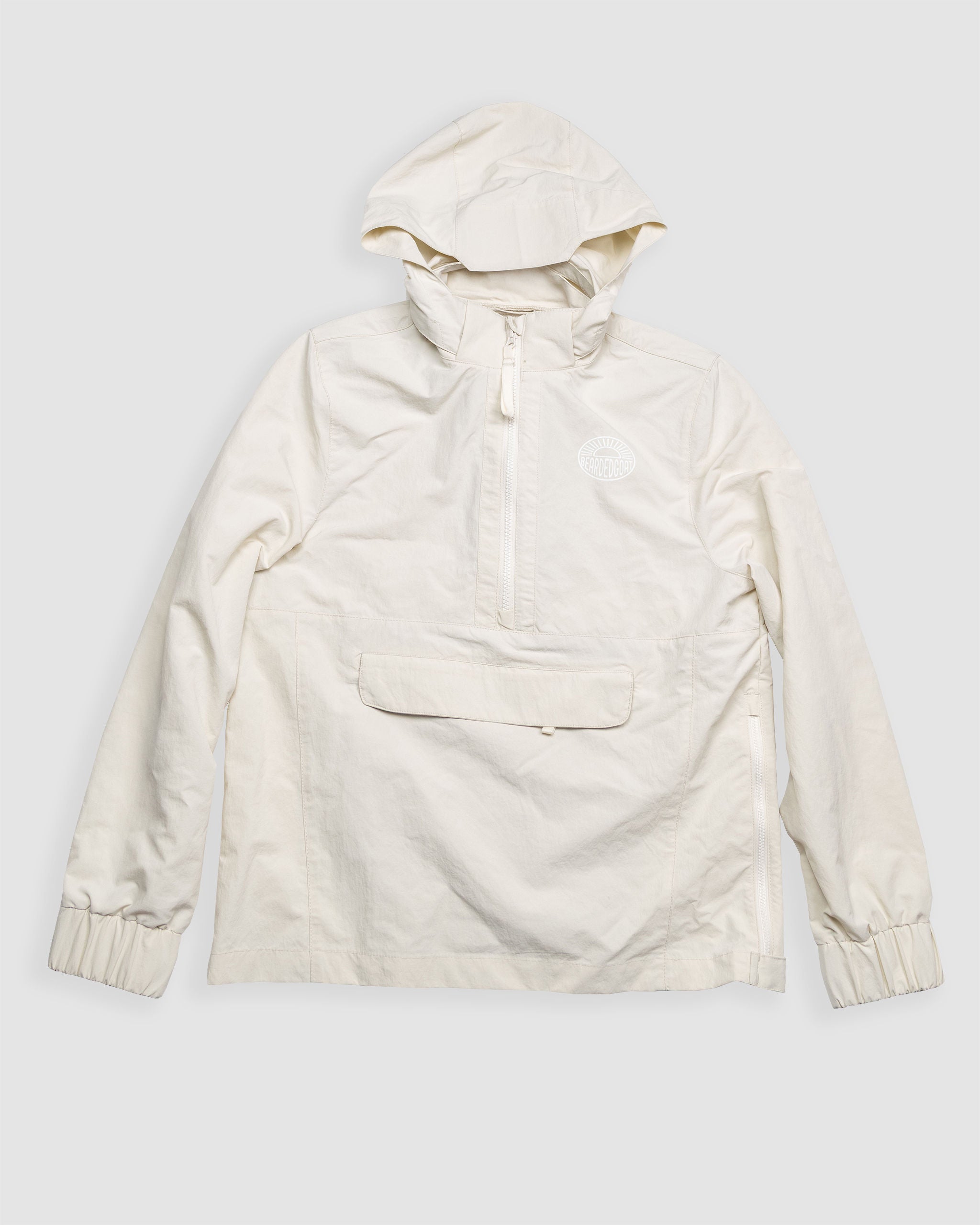 The BEARDEDGOAT Womens Deluge Pullover is a light beige, water-resistant hooded jacket with a front zipper, large flap pocket, elastic cuffs, and small circular chest logo, shown laid flat on a plain white background.