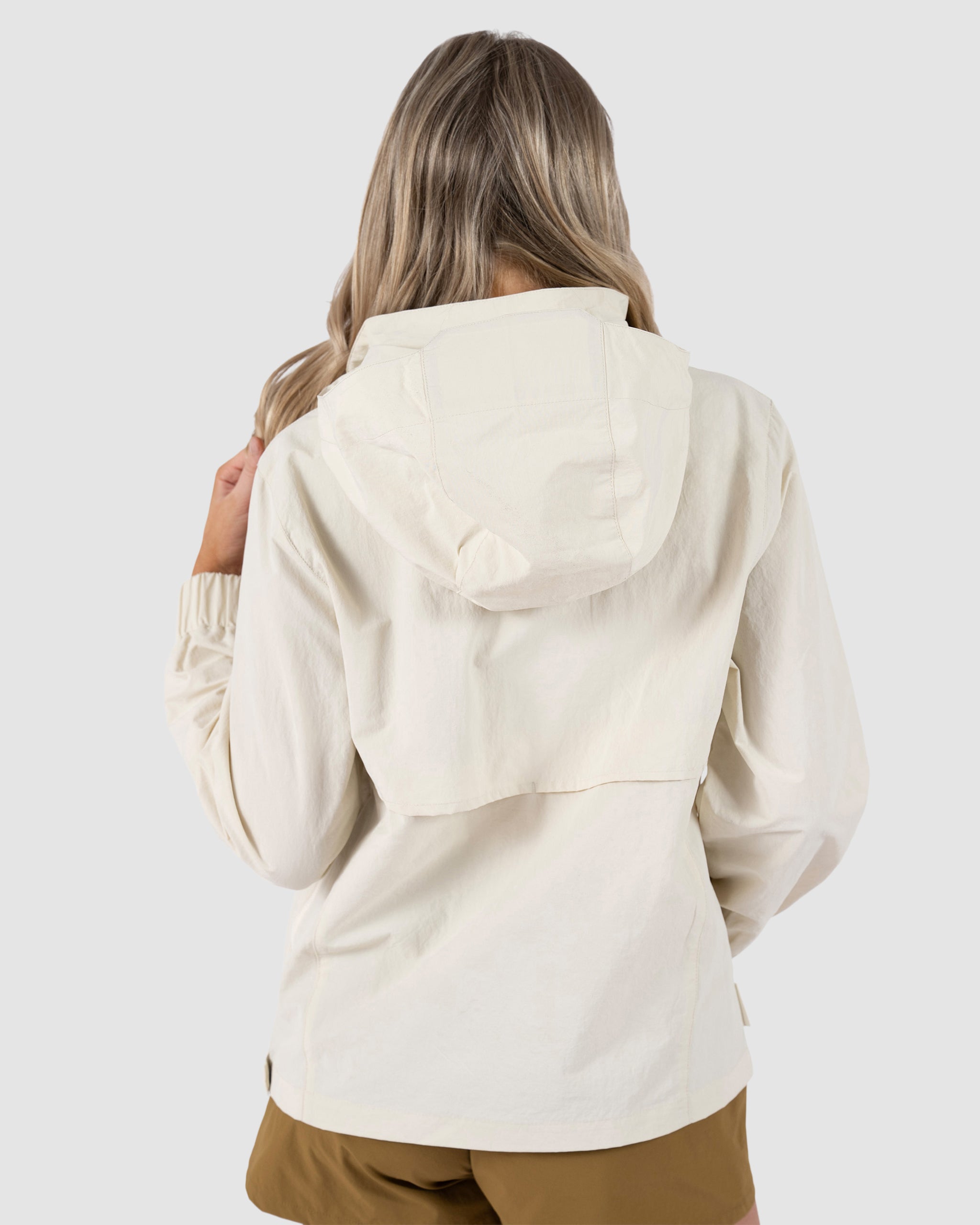 Seen from behind against a light gray background, a person with long, wavy blonde hair wears the BEARDEDGOAT Womens Deluge Pullover in cream with removable hood and brown shorts.