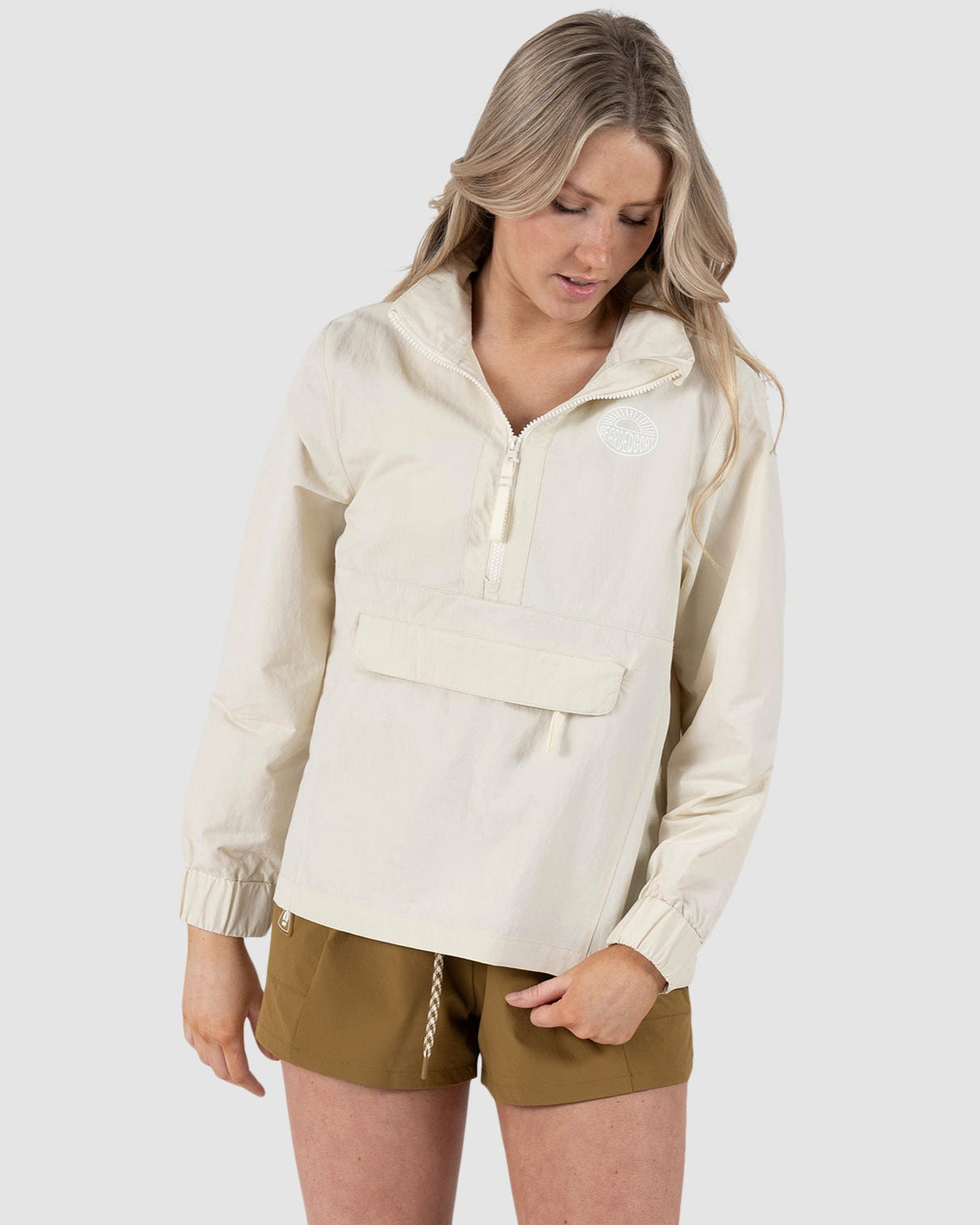 A woman with long blonde hair wears the BEARDEDGOAT Womens Deluge Pullover in cream, paired with brown shorts and white sneakers, standing against a plain light gray background and looking down.