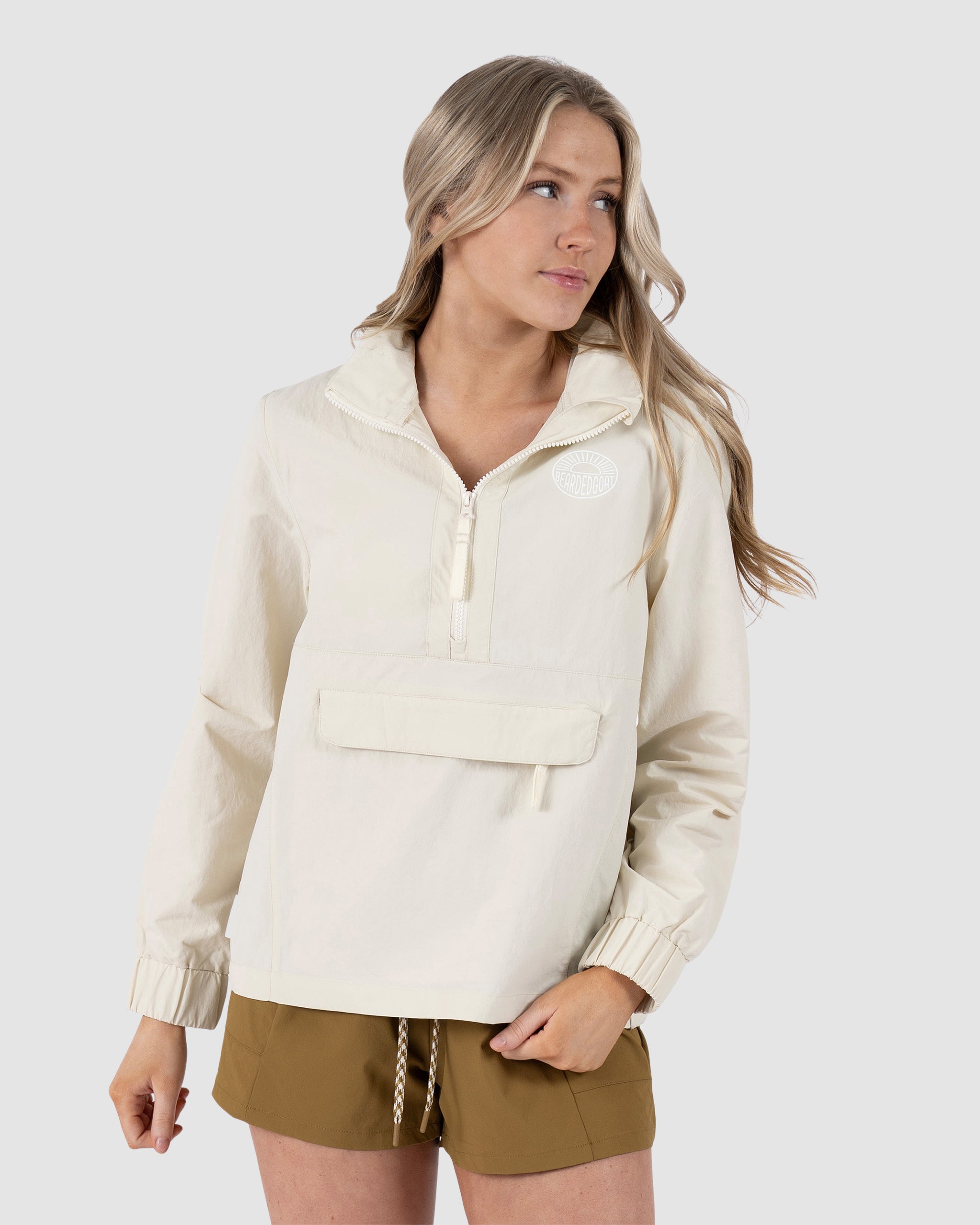 A woman with long blonde hair wears the BEARDEDGOAT Womens Deluge Pullover—a light beige, water-resistant zip-up jacket with a removable hood and front pocket—paired with brown shorts, standing against a plain light gray background.