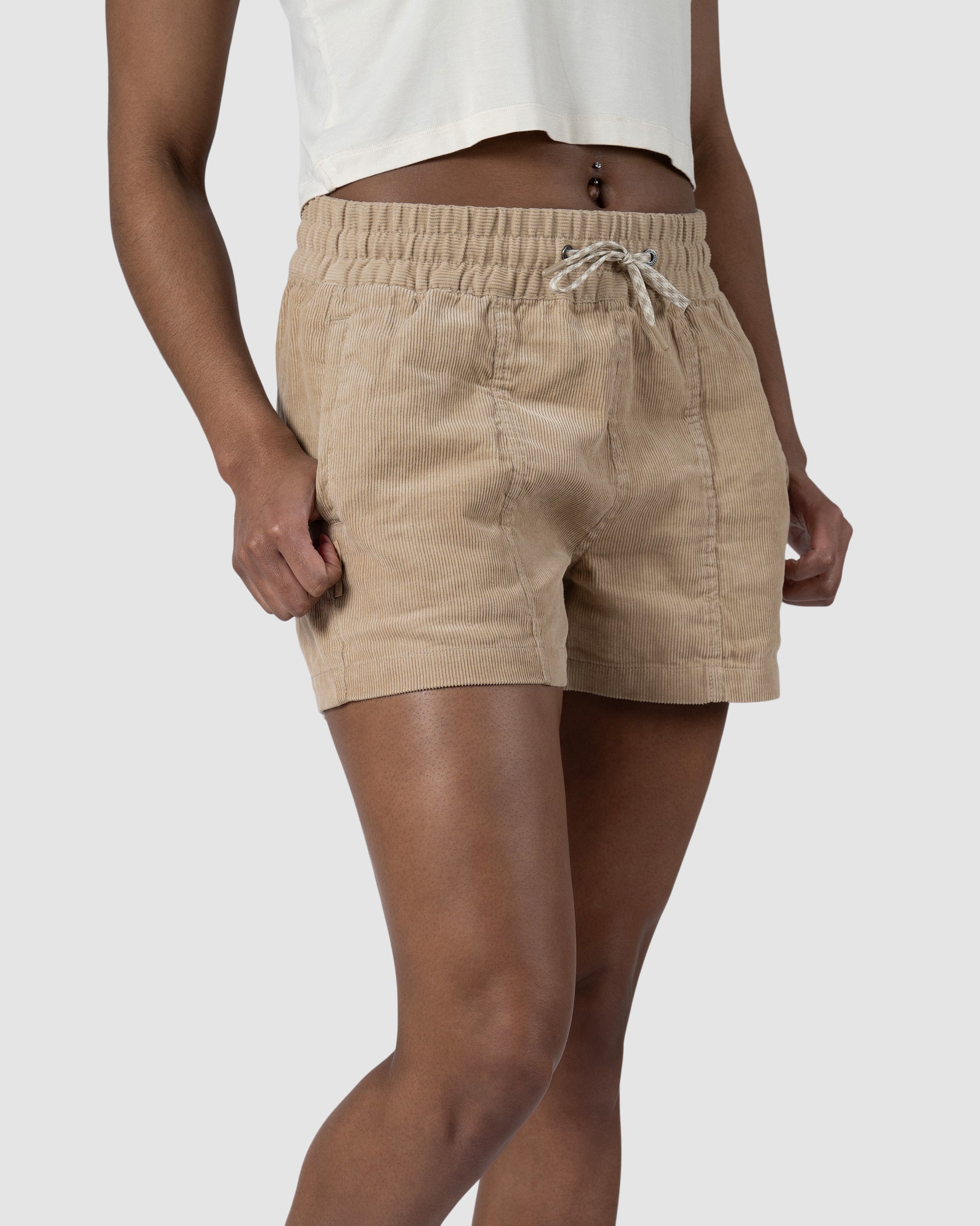 Model wearing BEARDEDGOAT Womens Cord Short in tan with an elastic waistband, paired with a white cropped top. Belly button piercing visible. Shot against a plain light gray background.