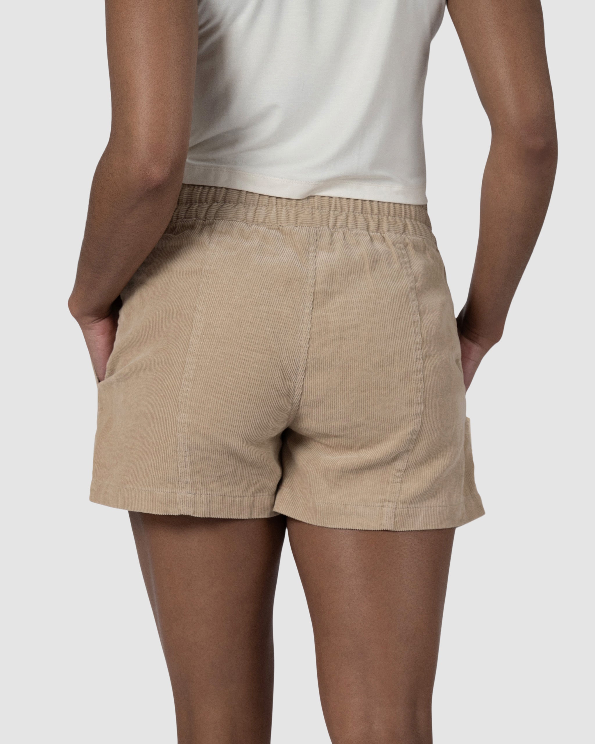 A person stands with hands in pockets, facing away from the camera against a gray background, wearing BEARDEDGOAT Womens Cord Short and a white shirt.