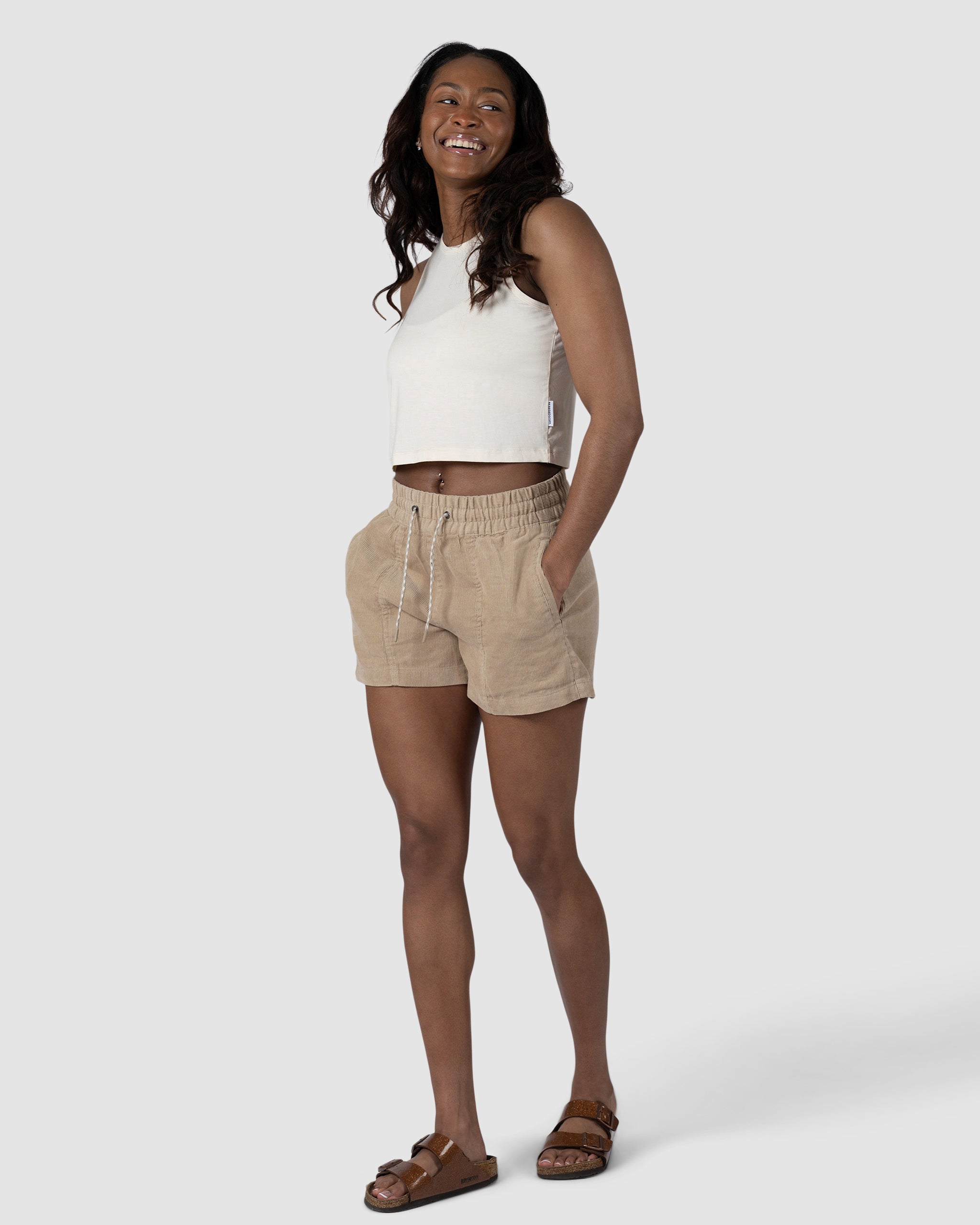 A woman smiles with her hands in her pockets, wearing BEARDEDGOATs Womens Cord Short—tan corduroy with an elastic waistband—paired with a white sleeveless top and brown sandals, posing against a light gray background.