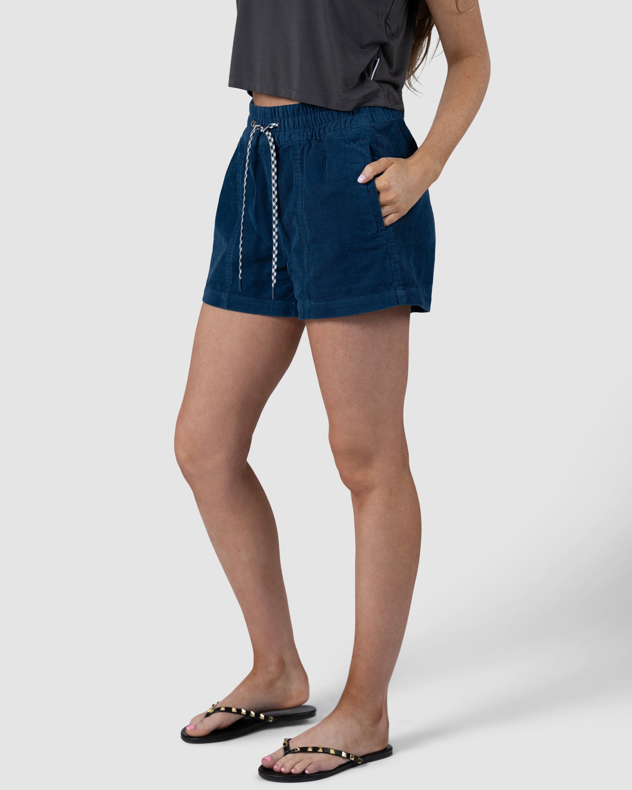 A woman stands with one hand in her pocket, wearing a dark gray top, BEARDEDGOAT Womens Cord Short in blue with an elastic waistband, and black sandals with gold studs against a plain light background.