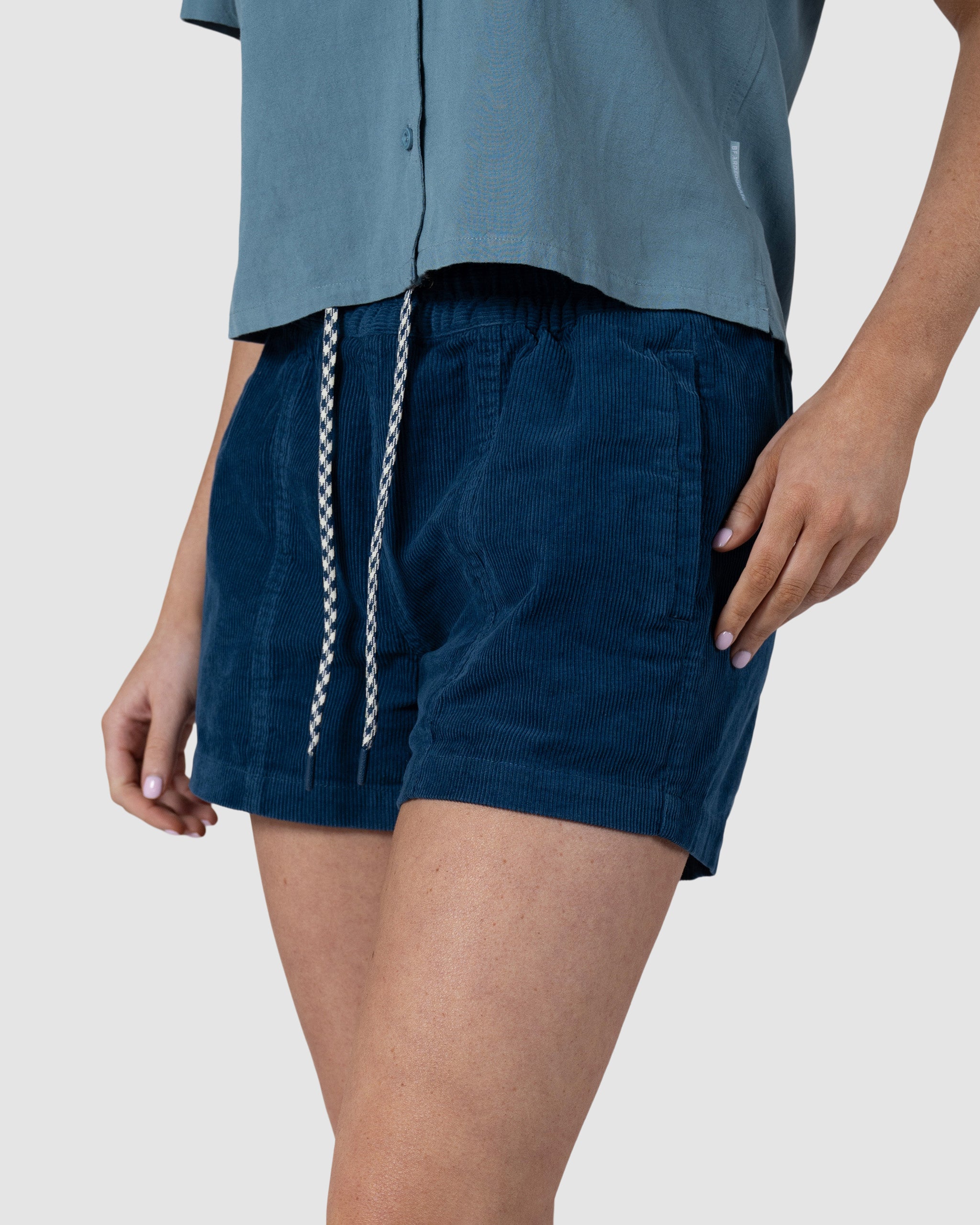 A person wears the BEARDEDGOAT Womens Cord Short in dark blue, styled with a light blue short-sleeved button-up shirt. The shot, set against a plain light background, is cropped to show only the torso and upper legs.