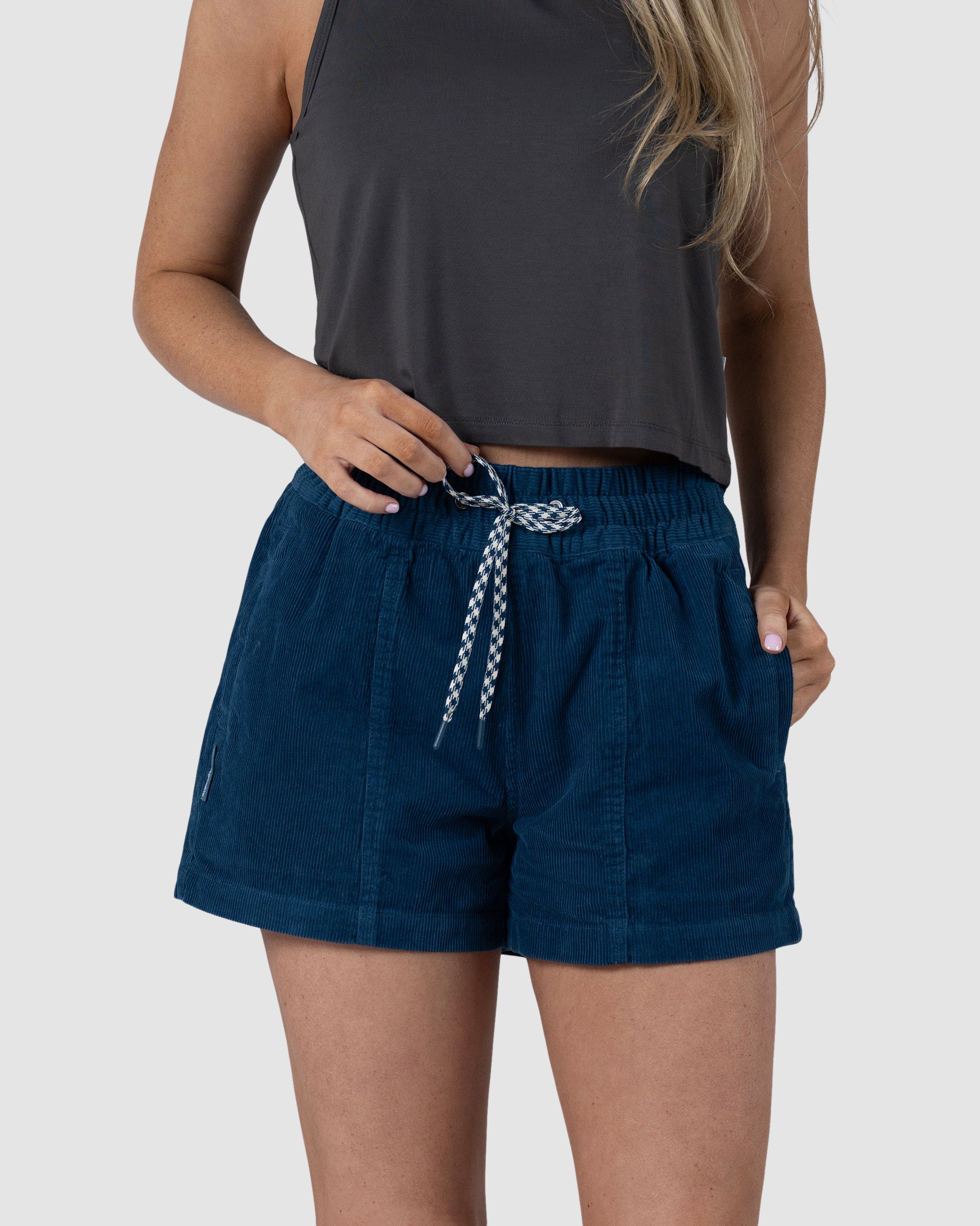 A woman wears the BEARDEDGOAT Womens Cord Short—a mid-rise blue style with an elastic waistband—paired with a dark gray sleeveless top. She stands against a plain light background, holding the drawstring and resting her left hand in her pocket.