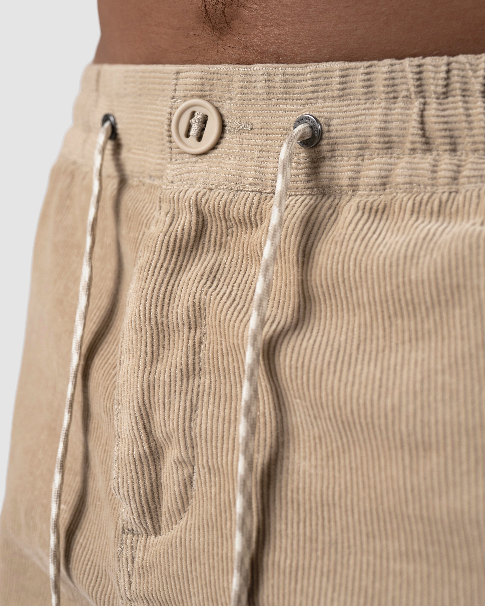 Close-up of a person wearing BEARDEDGOAT Mens Cord Short in beige, featuring a vintage corduroy design with a waistband button and white patterned drawstrings that add stylish contrast.