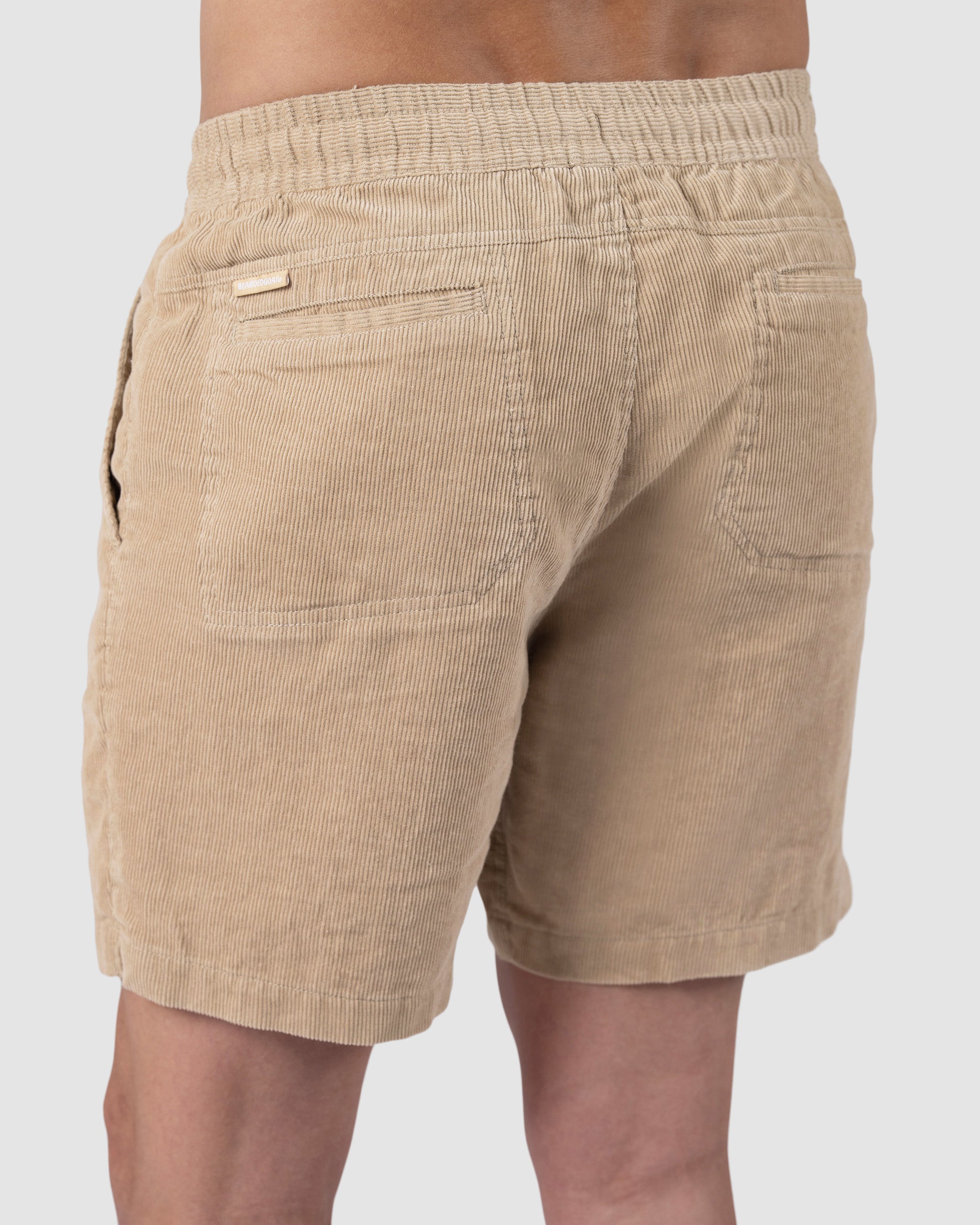 A person is seen from behind wearing BEARDEDGOAT Mens Cord Short in beige, featuring textured corduroy fabric, an elastic waistband, and two back pockets. The classic design offers vintage vibes against a light background.