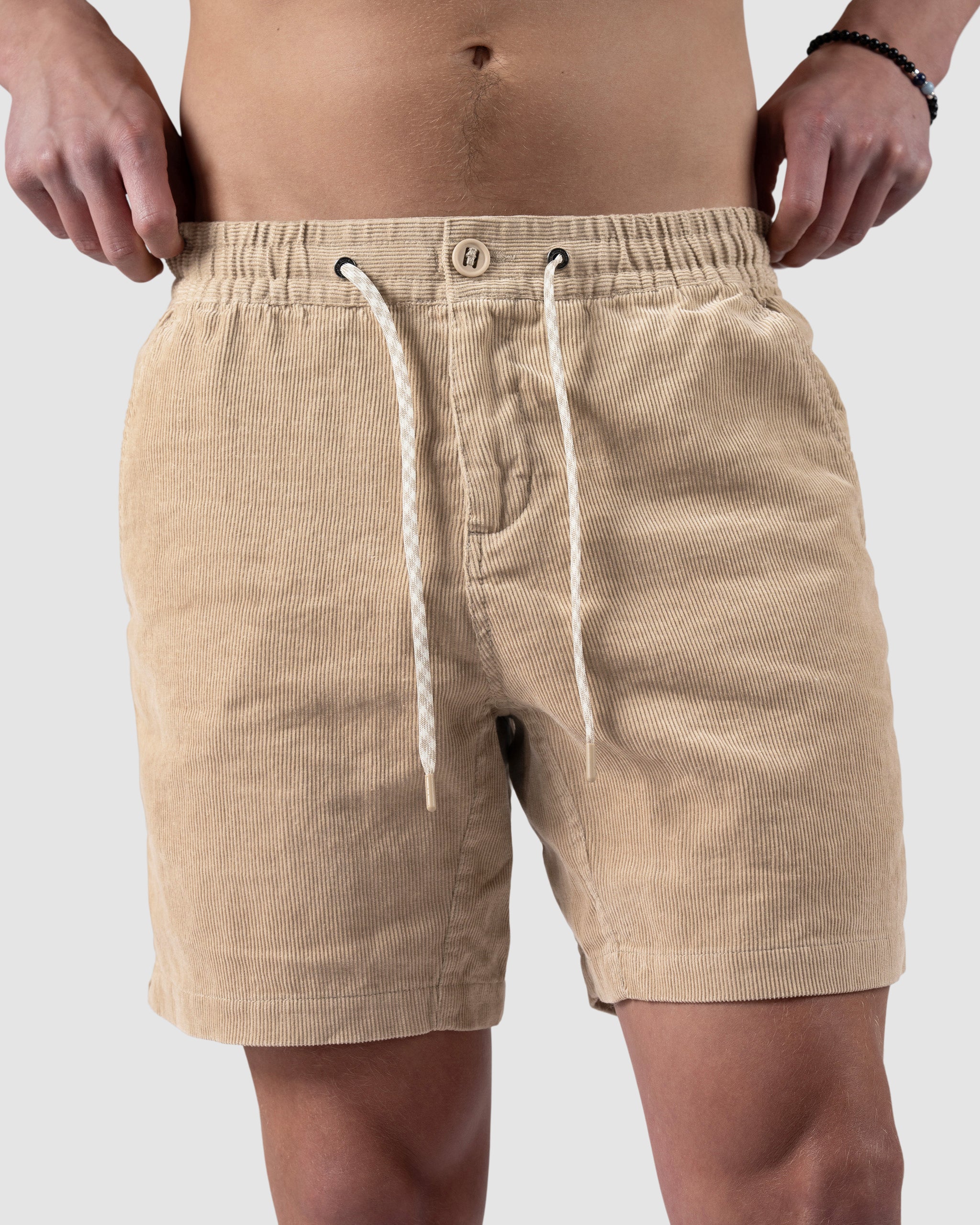A person showcases BEARDEDGOAT’s Mens Cord Short—tan corduroy with an elastic waistband, white drawstrings, and side pockets—holding the waistband to reveal part of their torso, evoking vintage vibes against a plain background.