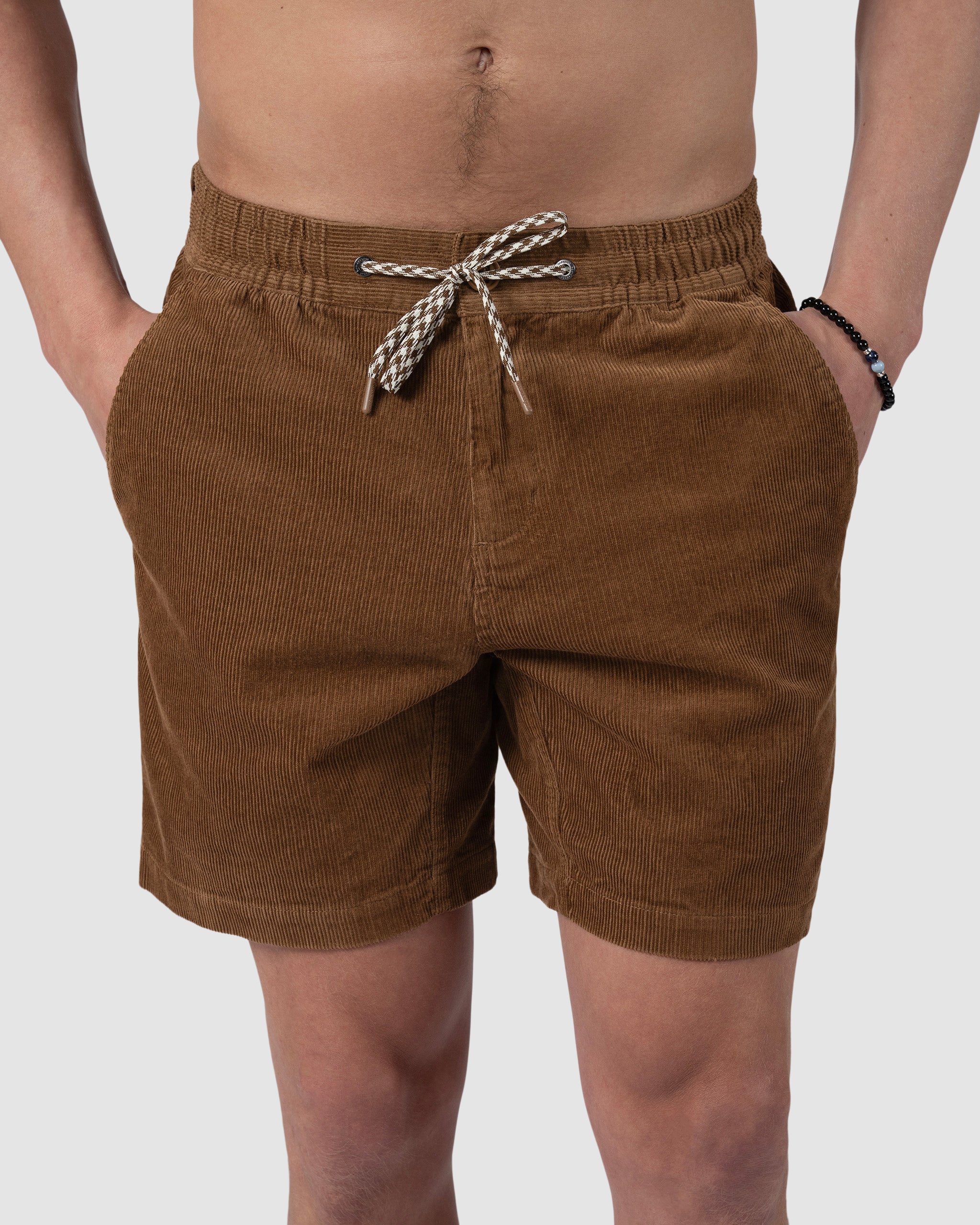 Wearing BEARDEDGOAT’s Mens Cord Short—brown corduroy with an elastic waistband and black-and-white drawstring—a shirtless individual, hands in pockets and a beaded bracelet on one wrist, radiates vintage summer style.