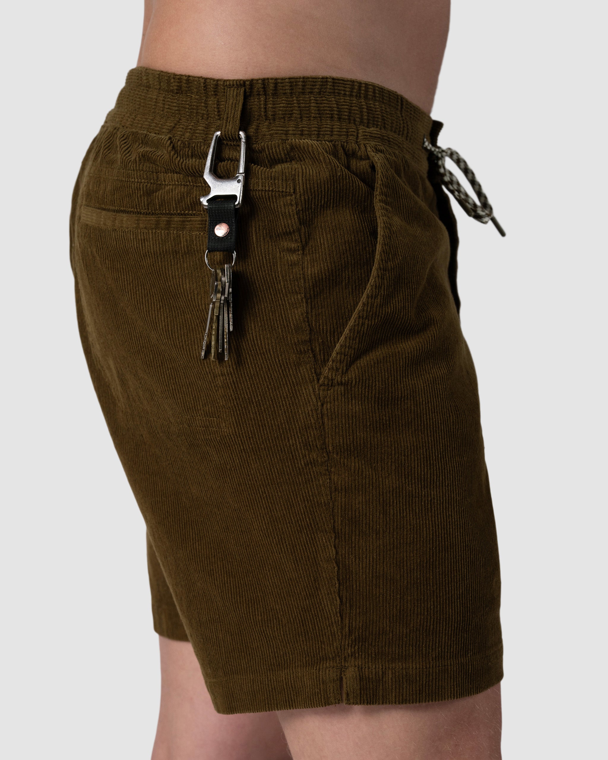 A person wearing BEARDEDGOAT Mens Cord Short in olive green, featuring an elastic waistband, braided drawstring, side pockets, and a metal clasp keychain on the belt loop for subtle vintage vibes.