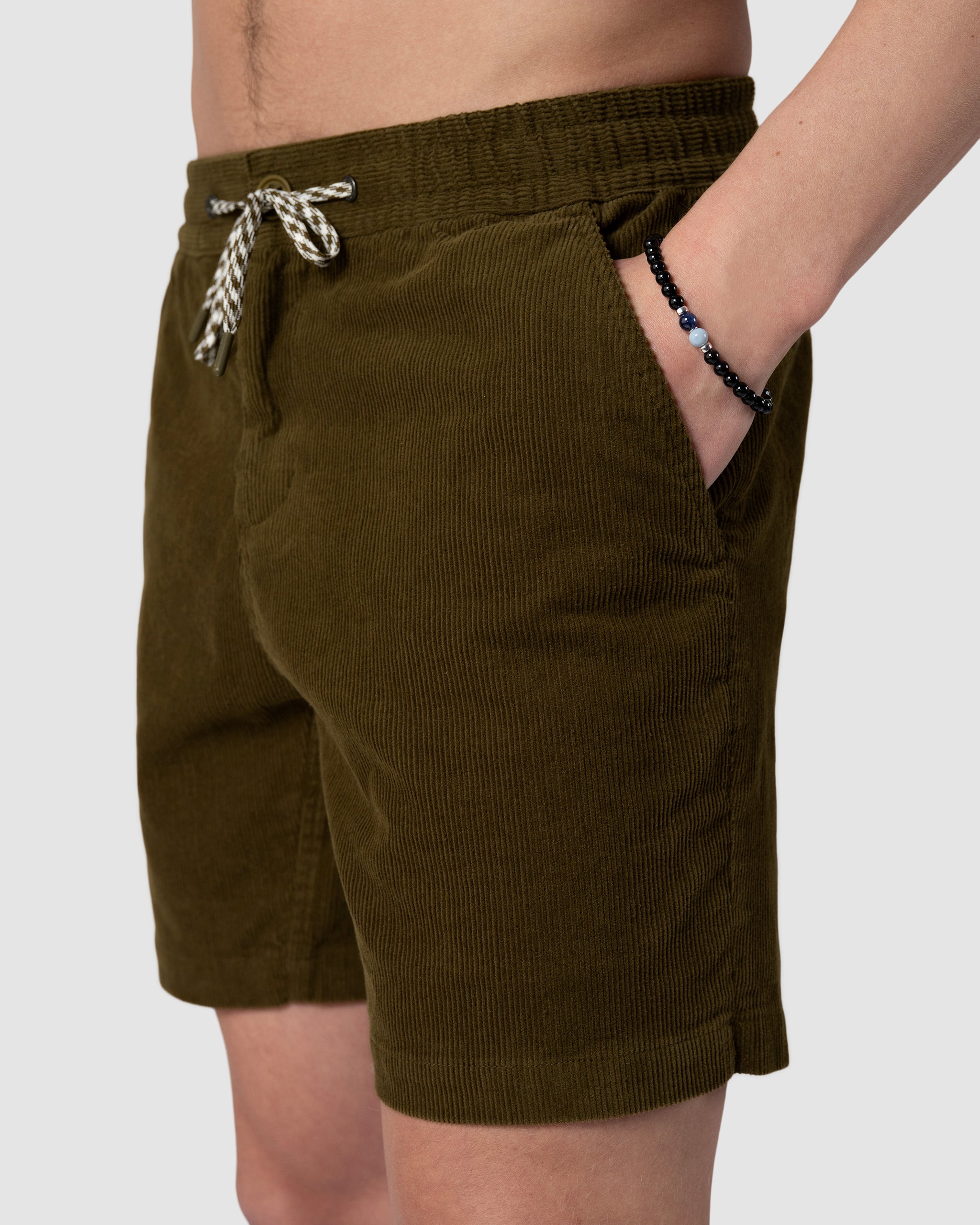 Wearing BEARDEDGOAT Mens Cord Short in olive green with a drawstring waist and pockets, this look showcases a black beaded bracelet and evokes vintage vibes against a simple, light backdrop.