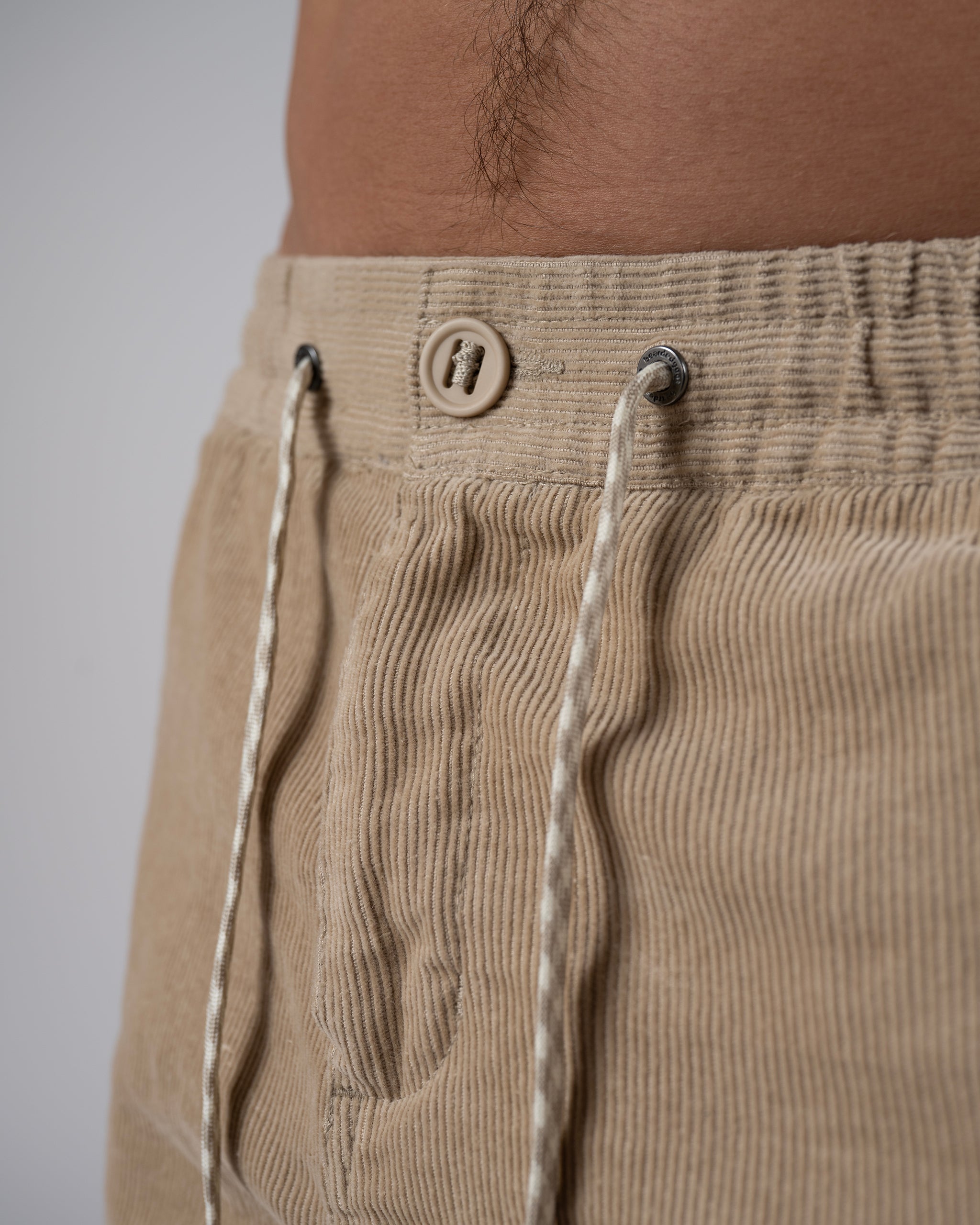 Close-up of a person wearing BEARDEDGOAT Men's Cord Short in beige, featuring a button and white drawstrings, showcasing their versatile everyday style and part of the lower abdomen.