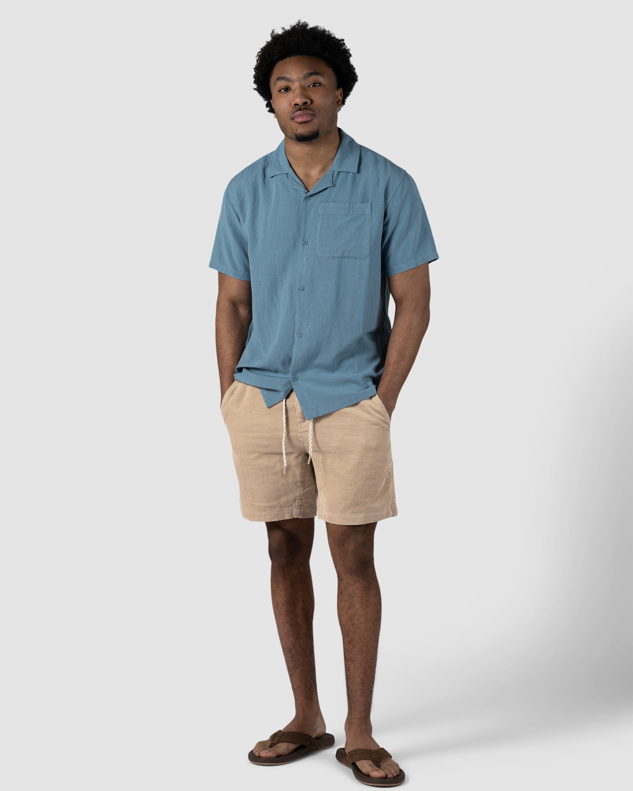 A man stands against a plain background wearing BEARDEDGOAT Mens Cord Shorts in tan with a drawstring, a light blue short-sleeve button-up shirt, and brown flip-flops. He has his hands in his pockets and looks relaxed.