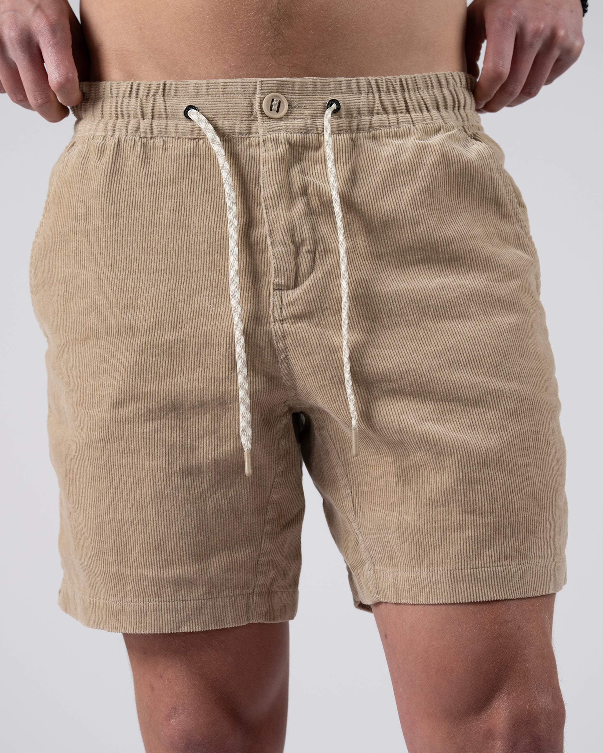 A man wears BEARDEDGOAT's Men's Cord Short—vintage corduroy with an elastic waistband, white drawstrings, button, and side pockets. The torso-to-mid-thigh crop highlights the shorts’ stylish everyday versatility.