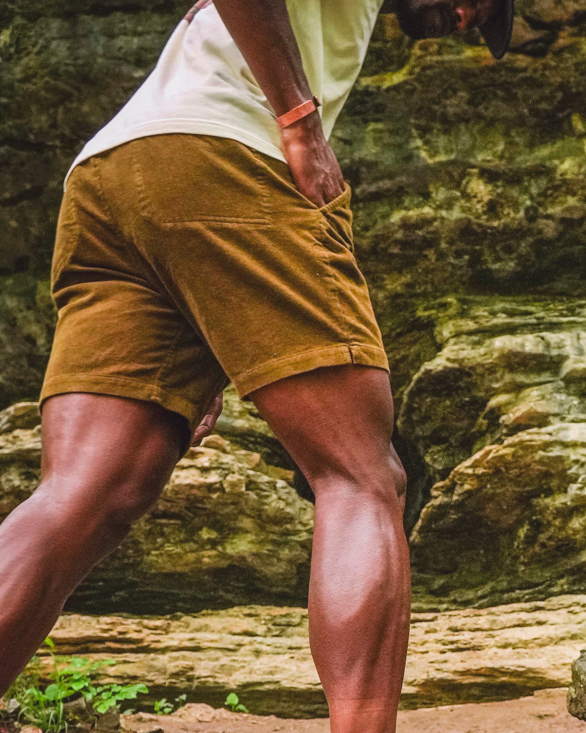 Wearing BEARDEDGOAT Men's Cord Short, a person stands outdoors on rocky terrain with one hand in their pocket, highlighting casual versatility against rough, layered rocks and greenery in the background.