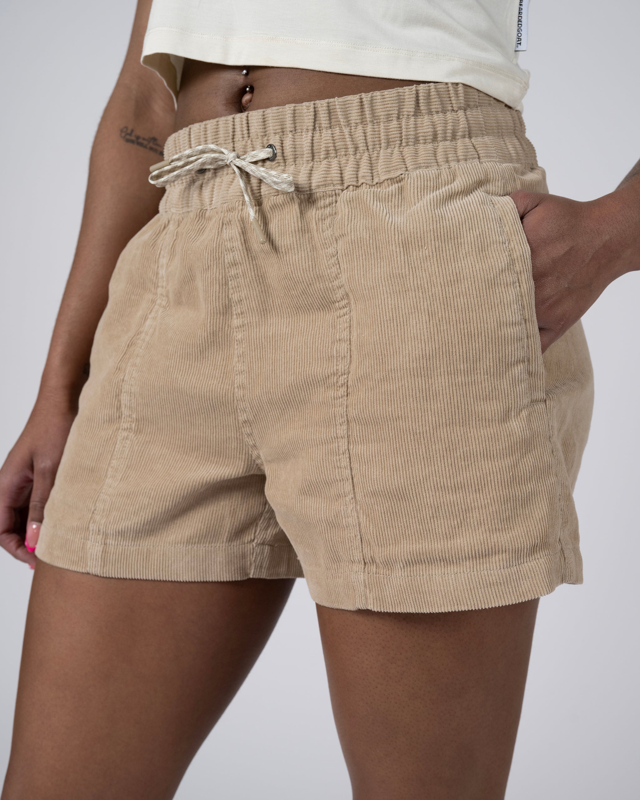 Model wears BEARDEDGOAT Women's Cord Short in beige, featuring an elastic waistband with drawstring and mid-rise fit. Styled with a light cropped top, one hand tucked in the pocket, showing lower torso and upper legs.