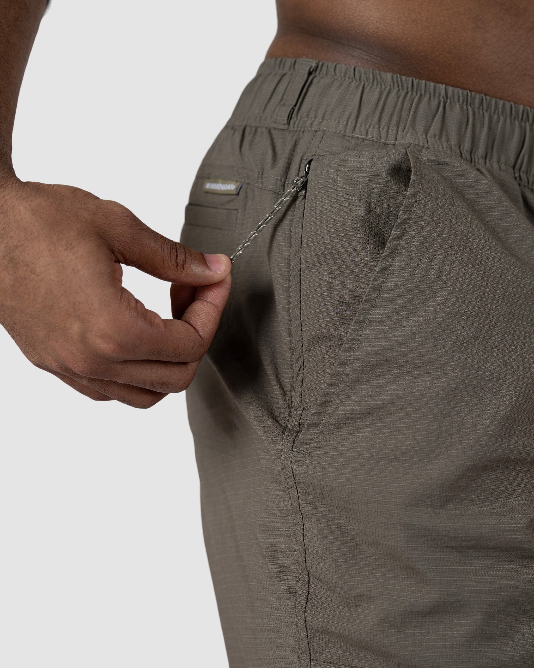 A person unzips the side pocket of BEARDEDGOAT Mens Canopy Short in olive green, made from lightweight stretch cotton ripstop, highlighting a close-up of the fabric and zipper detail.