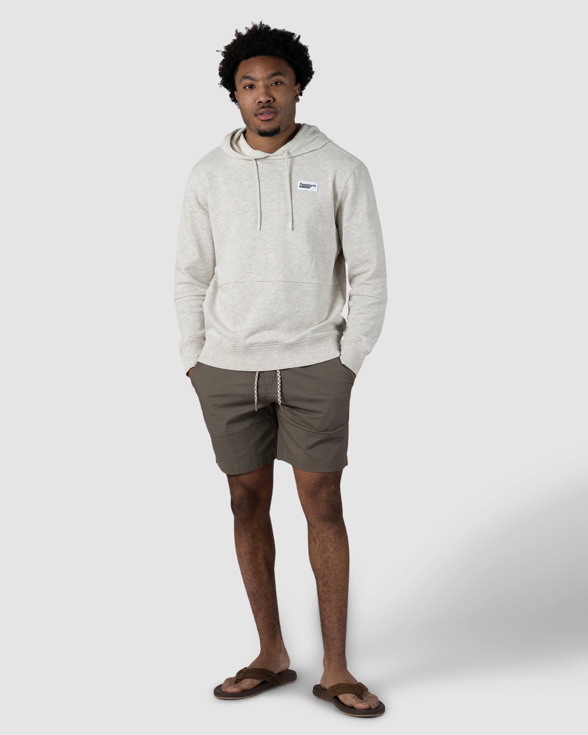 A man with short curly hair stands against a plain background wearing a light gray hoodie, BEARDEDGOAT Mens Canopy Short in olive green stretch cotton ripstop, and brown flip-flops, with his hands in his pockets.