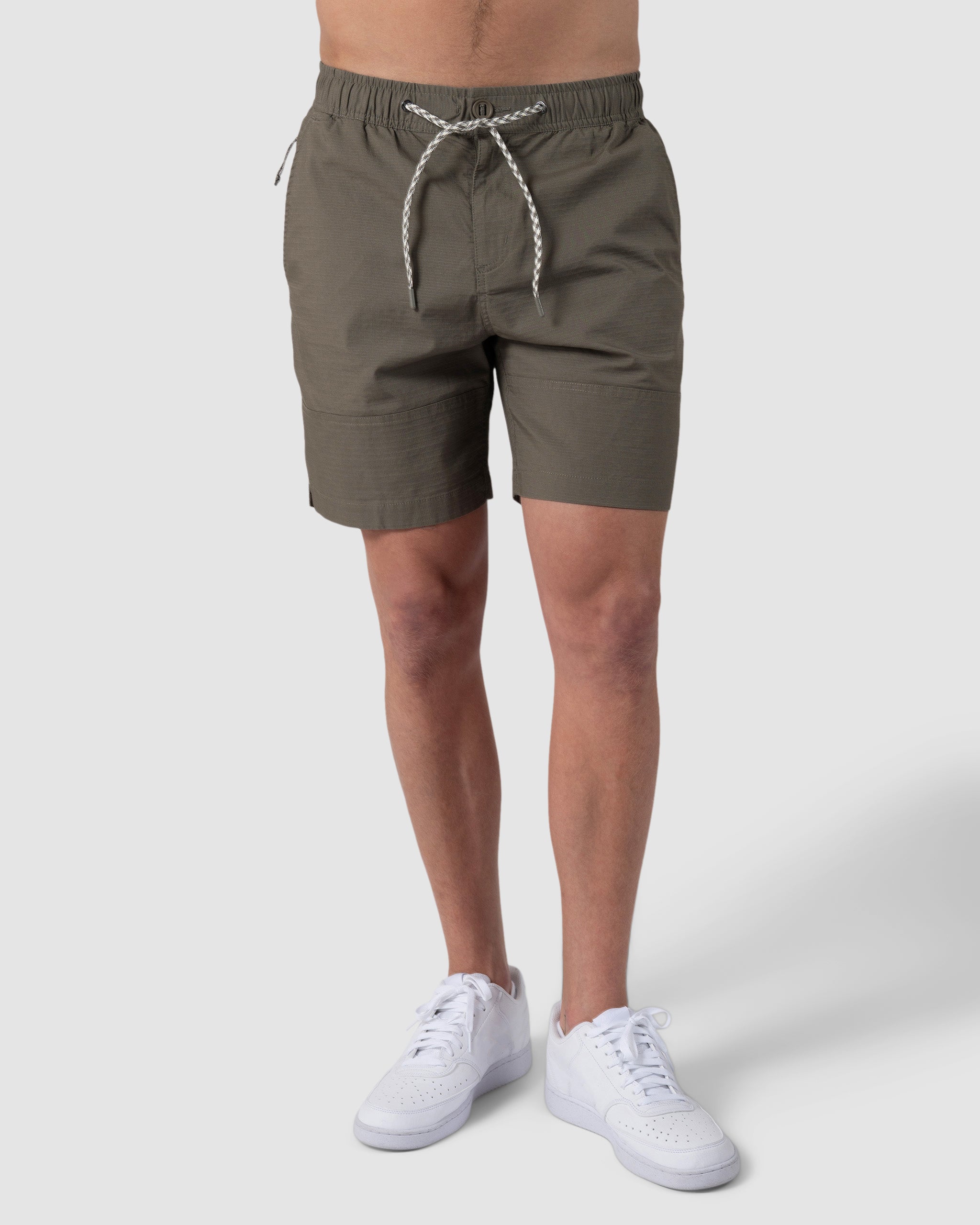 A person wearing BEARDEDGOAT Mens Canopy Short in olive green stretch cotton ripstop with white sneakers, standing against a plain light grey background.