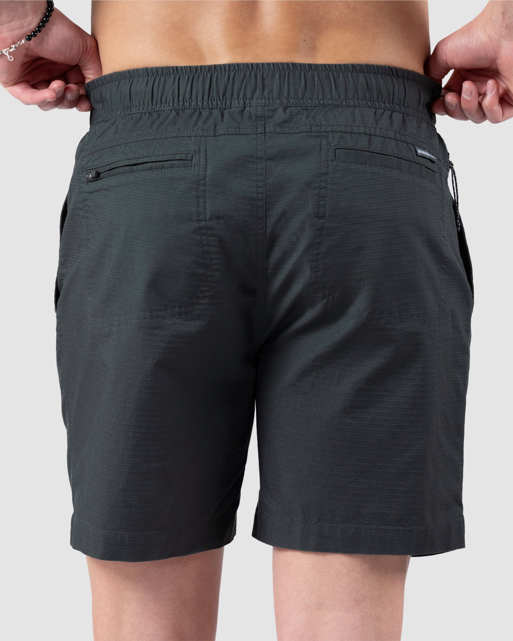 A man is seen from behind wearing BEARDEDGOAT Mens Canopy Shorts in dark green, made of lightweight stretch cotton ripstop with an elastic waistband and two zippered back pockets, set against a plain light gray background.