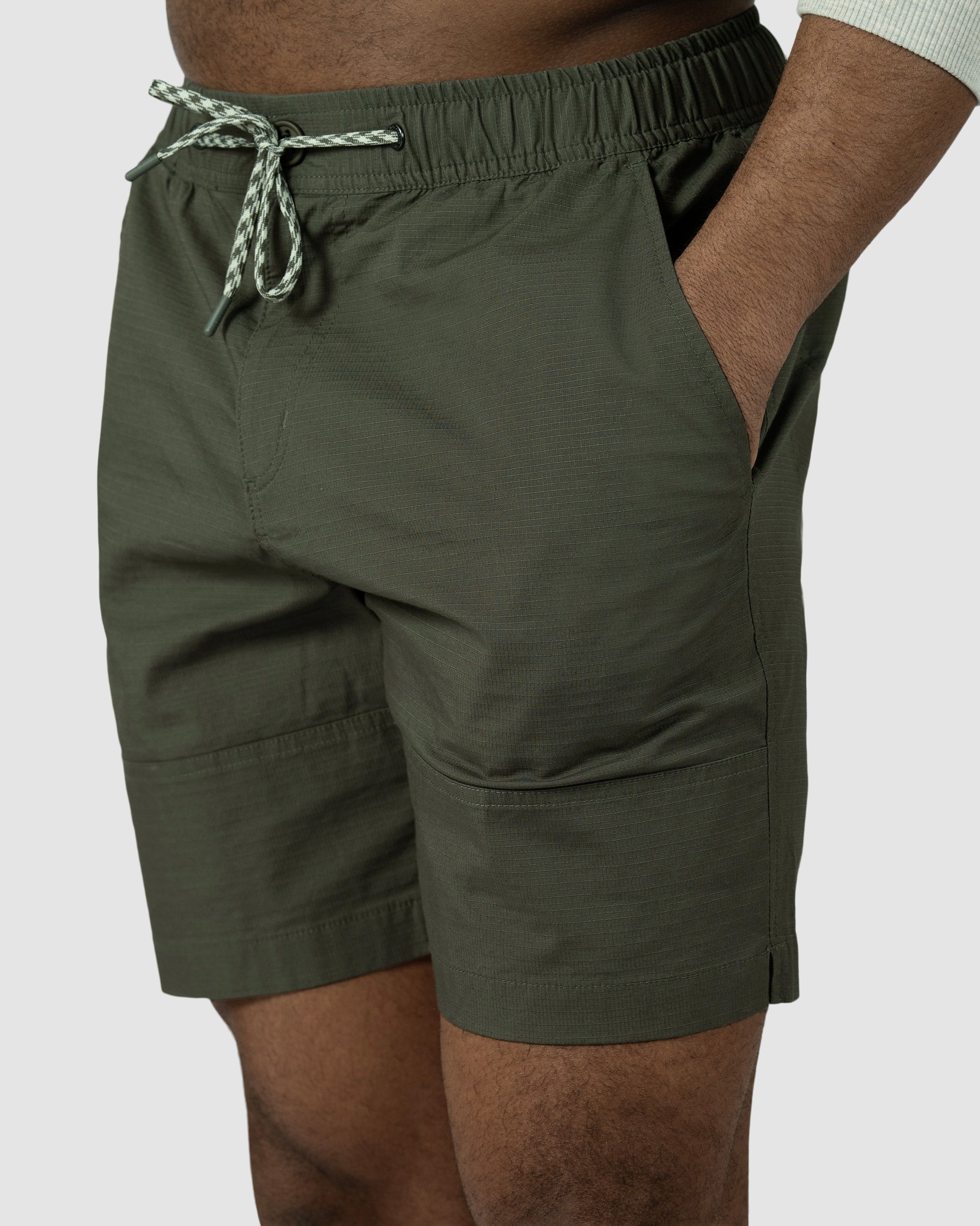 A man wears BEARDEDGOAT Mens Canopy Short in olive green, crafted from lightweight stretch cotton ripstop, hands in pockets, shown waist to mid-thigh against a light grey background.